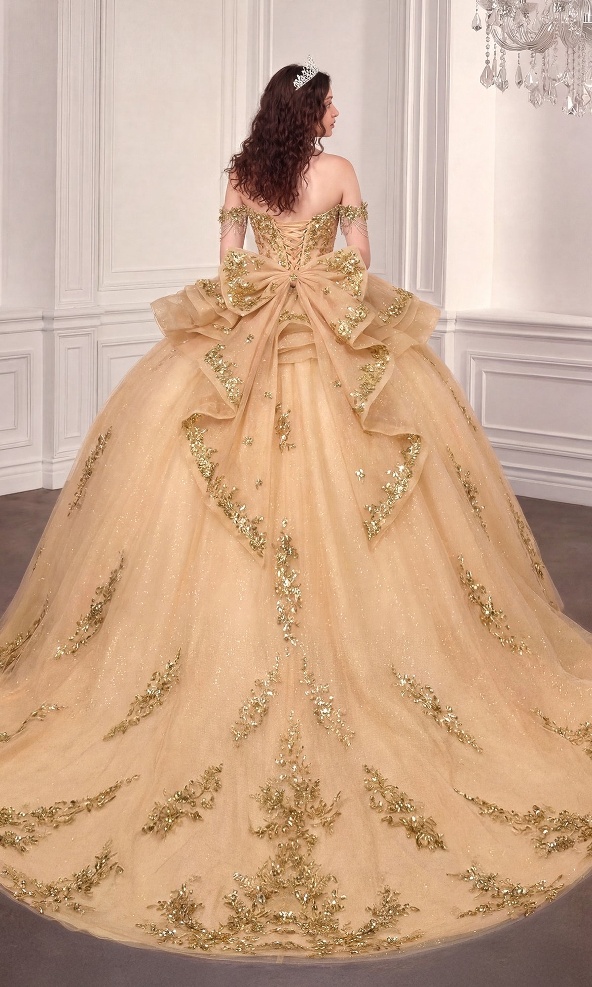 A woman stands gracefully in an elegant, light-colored room wearing the Quinceanera Dress 15746 By Ladivine—an off-the-shoulder ball gown featuring gold embellishments and a large bow on the back.