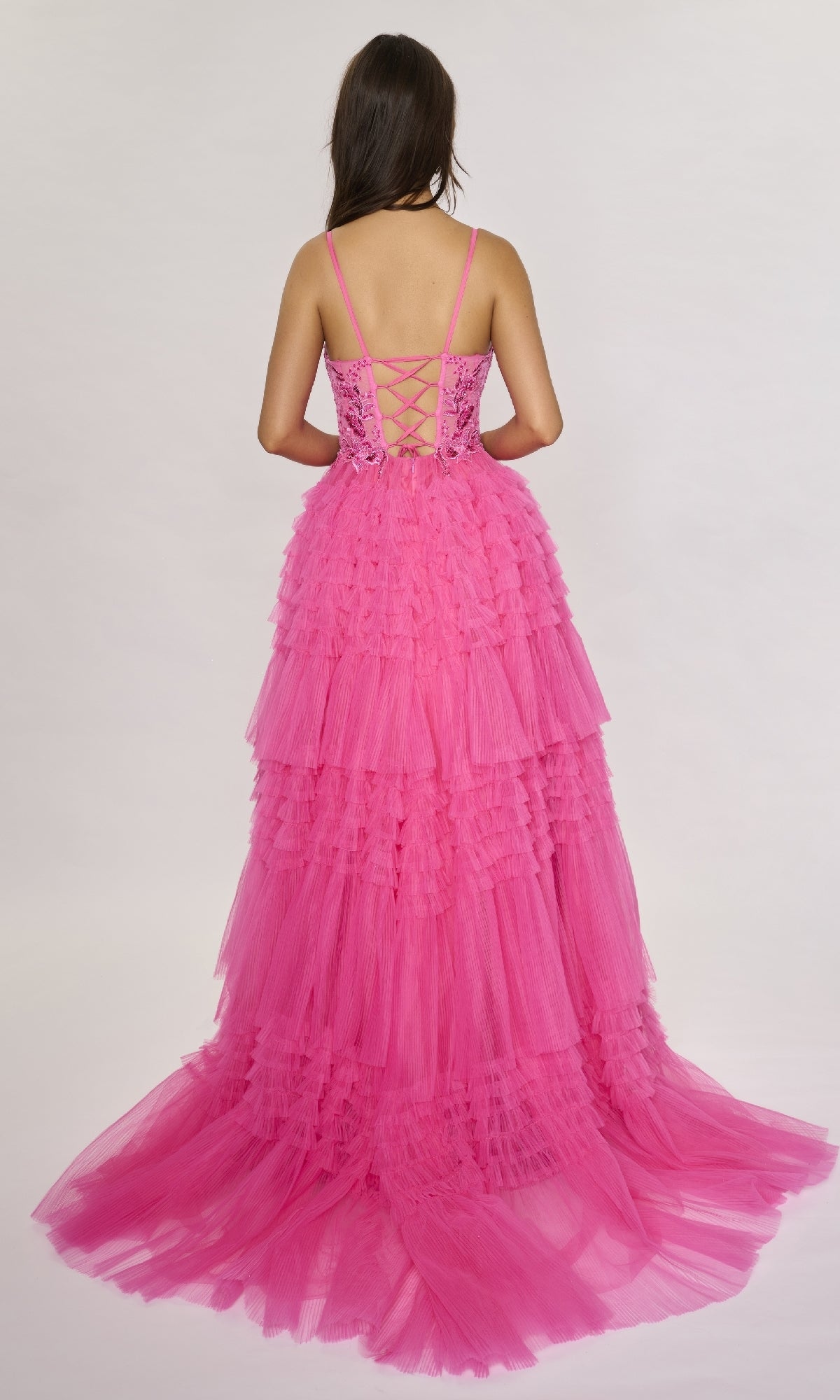 A woman with long brown hair faces away, wearing the Nina Canacci 1582—a bright pink long formal dress with a lace-up back and A-line ruffle tiered tulle skirt, flowing gracefully against a plain light background.