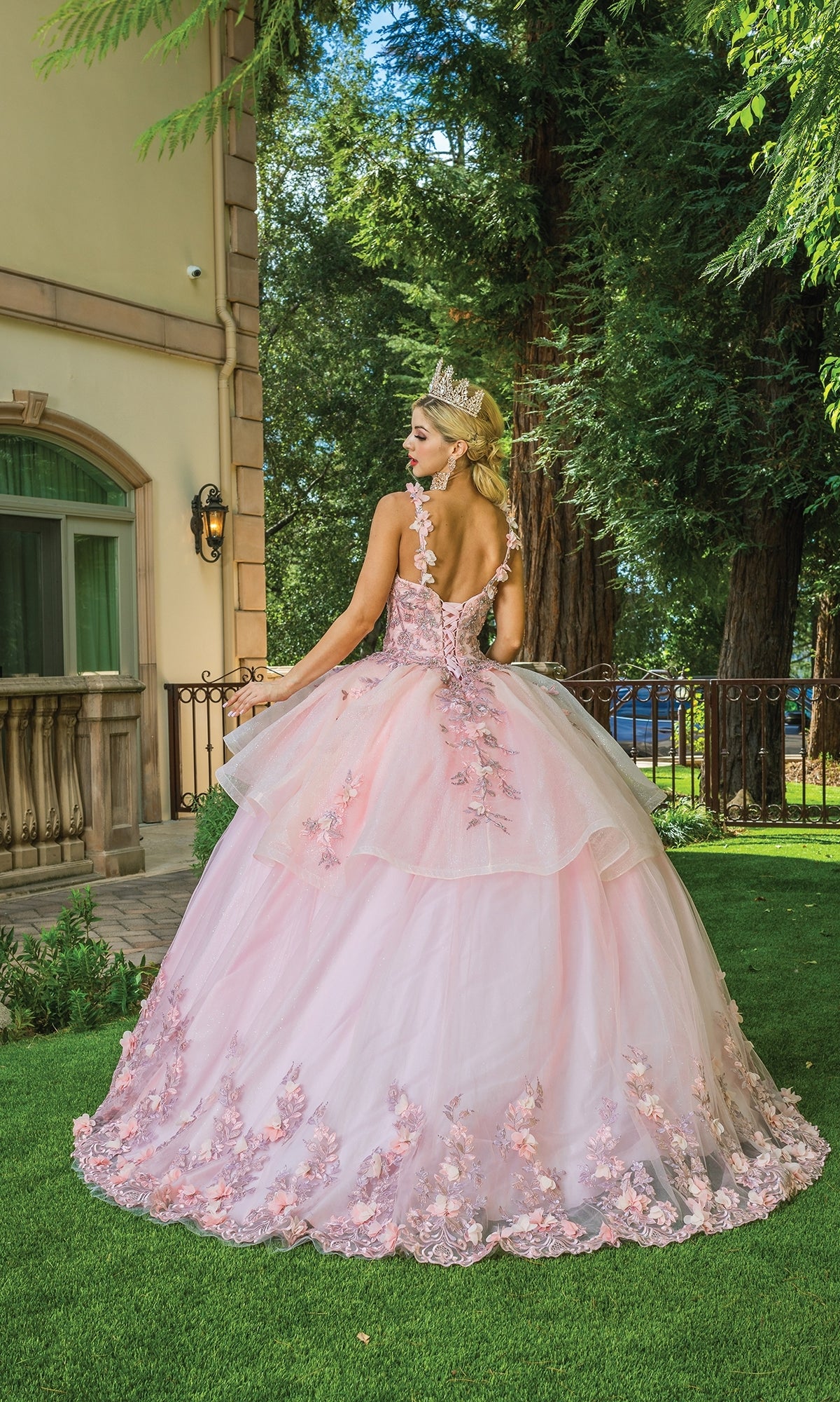 A woman in the Quinceanera Dress 1583 by Dancing Queen—a voluminous, light pink ball gown with floral details and a crown—stands on grass, facing away near a building and tall trees.