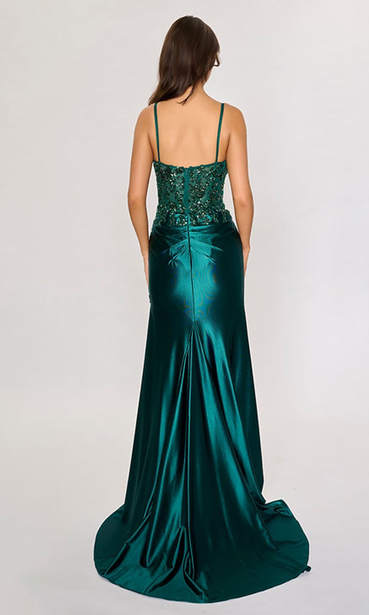 A woman stands with her back to the camera in a green Nina Canacci 1585 long formal dress featuring thin straps, a fitted bodice, and a slight train against a plain background.