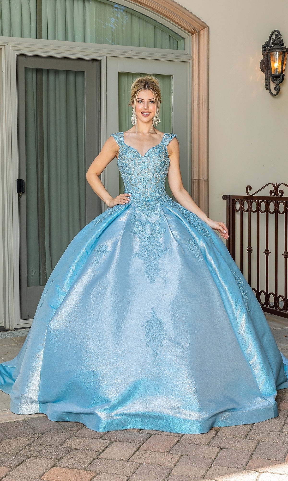 A woman stands outdoors by glass doors and a window, wearing the Quinceanera Dress 1585 by Dancing Queen—a light blue, floor-length ball gown with a fitted bodice, full skirt, and an elegant sweetheart neckline.