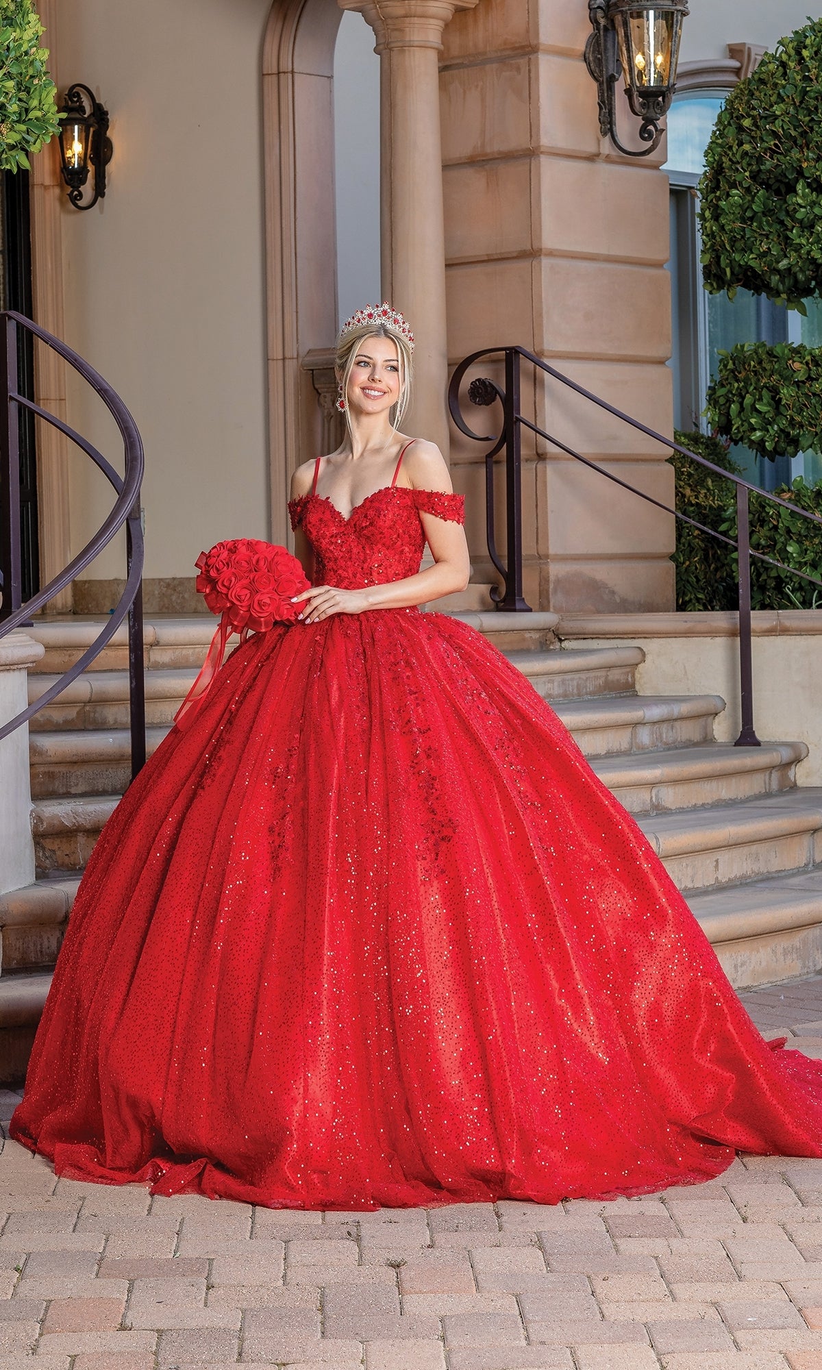 A young woman wearing the Quinceanera Dress 1595 by Dancing Queen stands on outdoor stairs, smiling at the camera with a tiara and a bouquet of red roses.