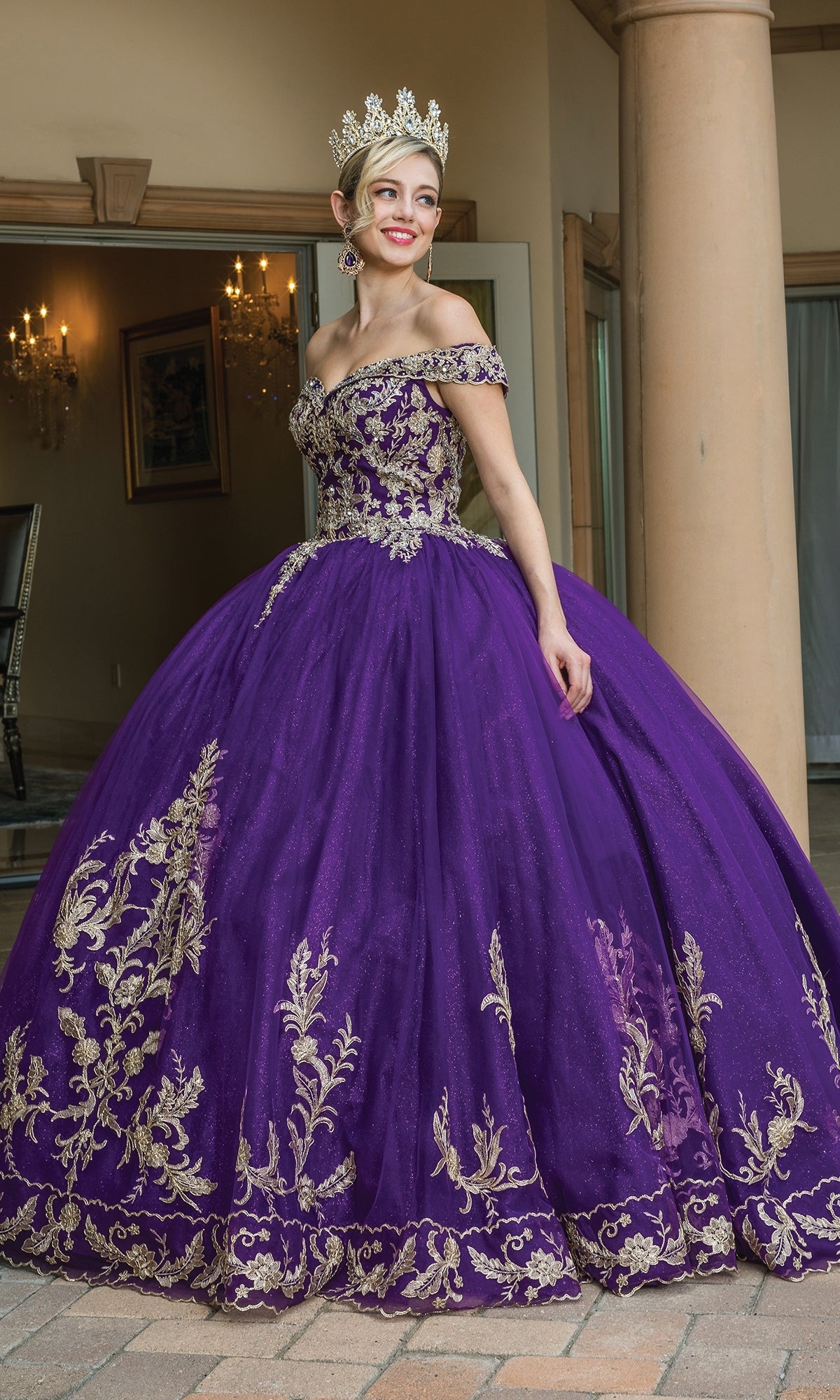 A woman stands on a tiled floor in front of a building entrance, wearing Quinceanera Dress 1596 by Dancing Queen, an off-the-shoulder purple a-line ball gown with gold embroidery and a gold crown.