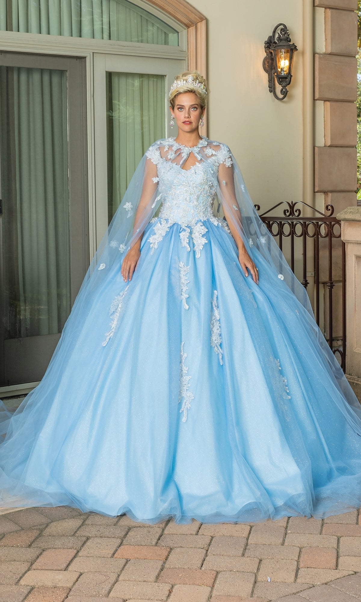 A person stands outside wearing the Quinceanera Dress 1597 By Dancing Queen, a light blue ball gown with floral lace embroidery, a sheer cape, and a floral headpiece.