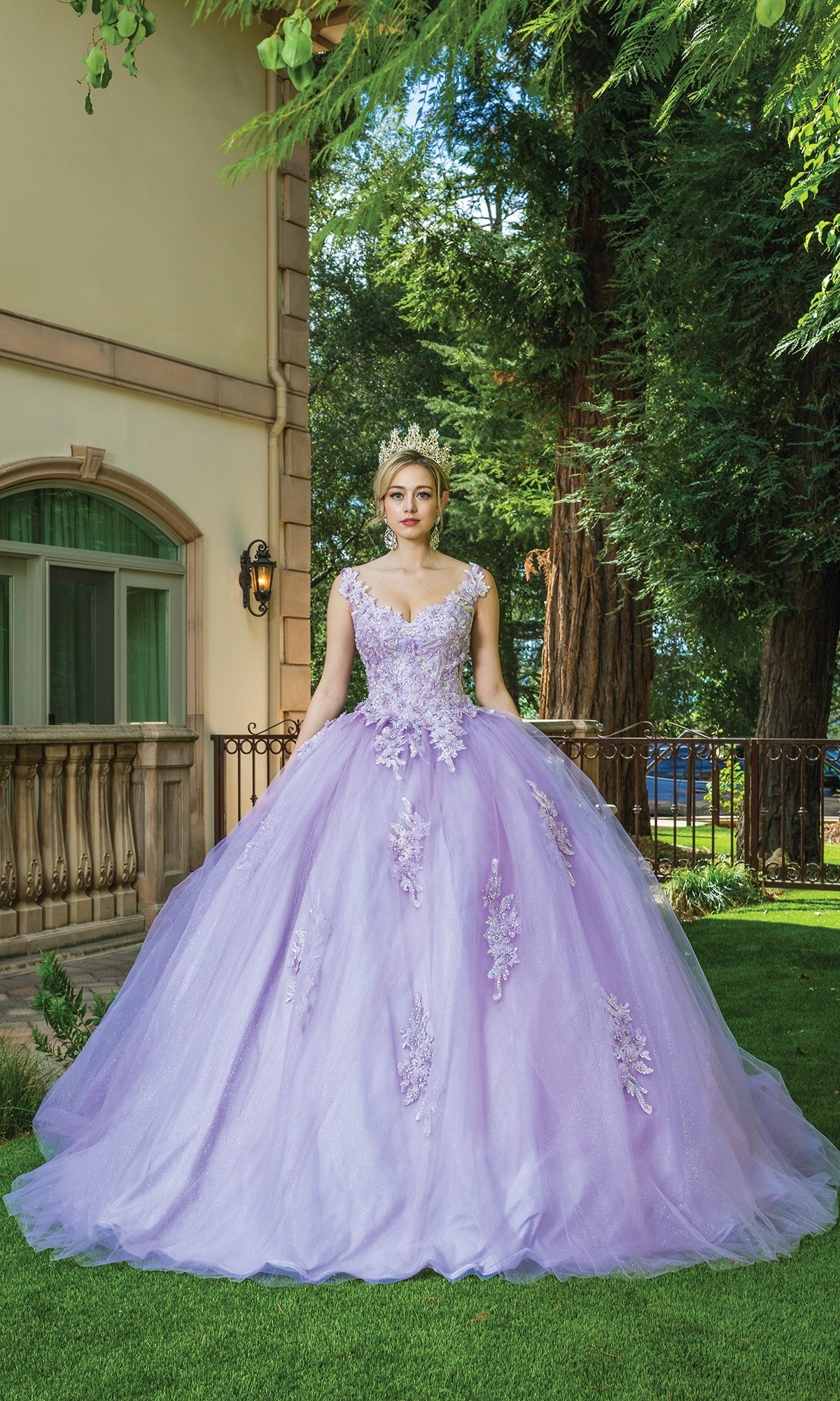 A woman in the Quinceanera Dress 1597 By Dancing Queen—a lavender ball gown with cap sleeves, floral details, and a tiara—stands on grass near a building and large trees.