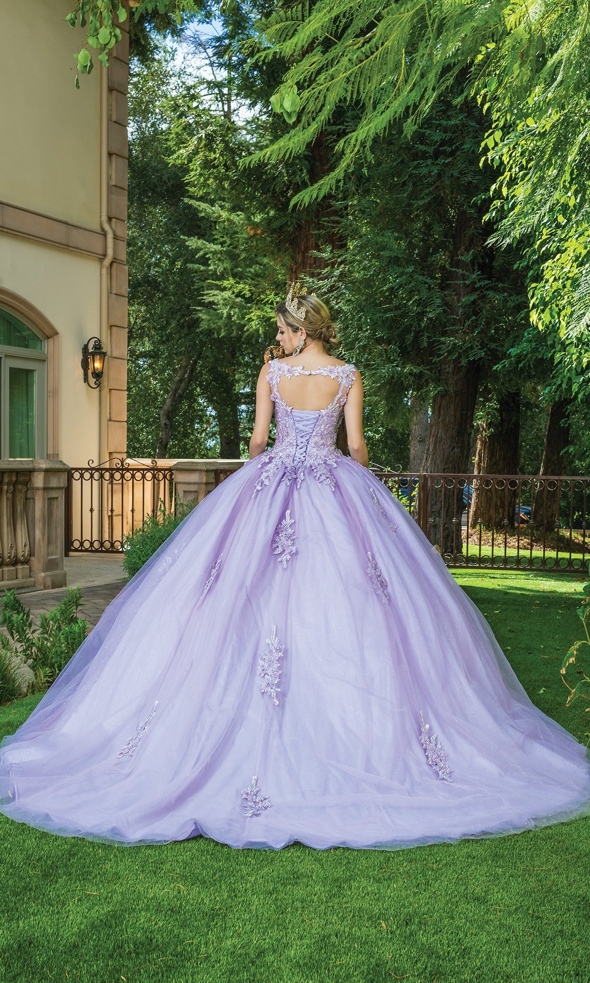 A woman stands on grass near a large house and trees, wearing the Quinceanera Dress 1597 By Dancing Queen—a lavender ball gown with cap sleeves and delicate lace embroidery—facing away outdoors.