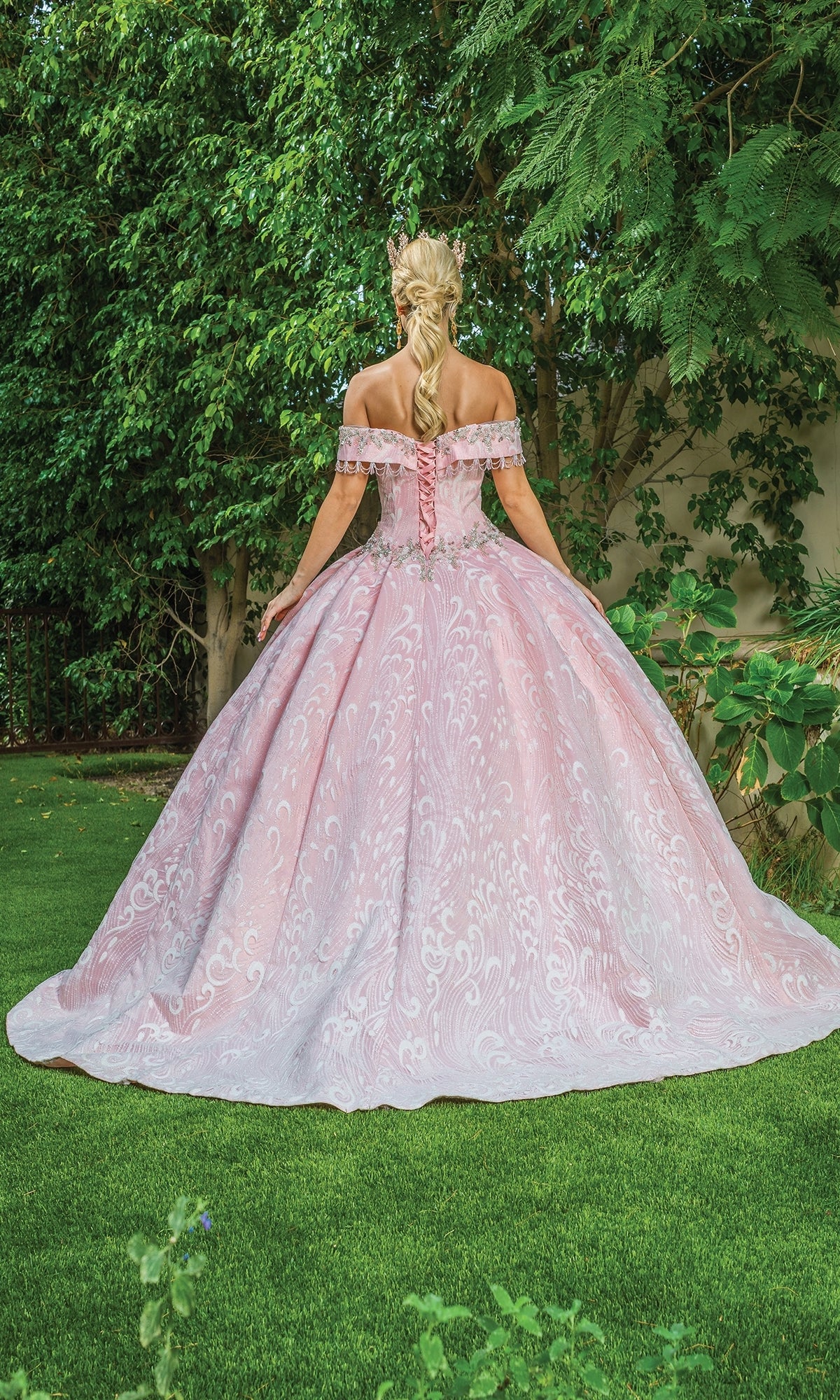 A woman with blonde braided hair stands on grass facing away, wearing the Pastel Pink Quinceanera Dress 1602—an off-the-shoulder blush ball gown—in front of green bushes.