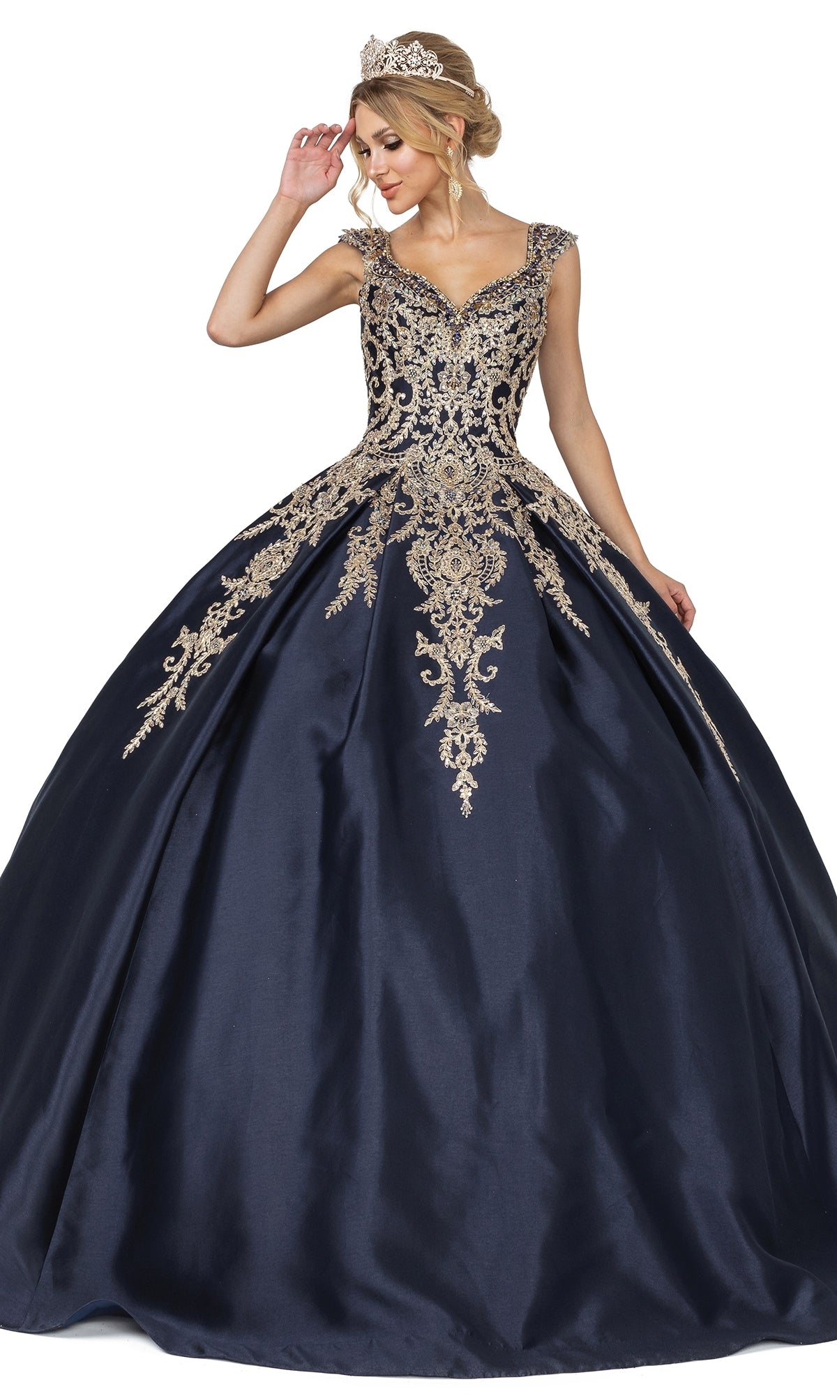 A woman wears the Quinceanera Dress 1606 By Dancing Queen, a navy blue gown with cap sleeves, gold embroidery, and a matching tiara, standing against a white background.