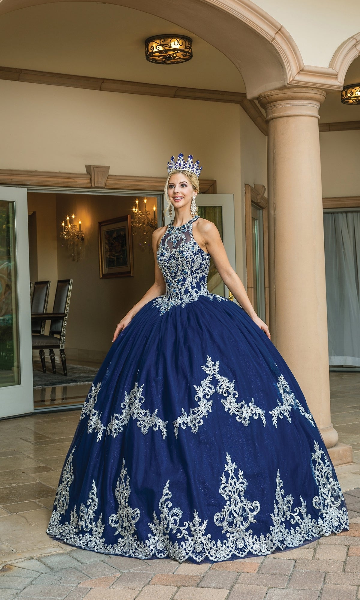A woman stands outside a building wearing the High-Neck Navy Quinceanera Dress 1607 with silver lace embroidery and a matching crown.