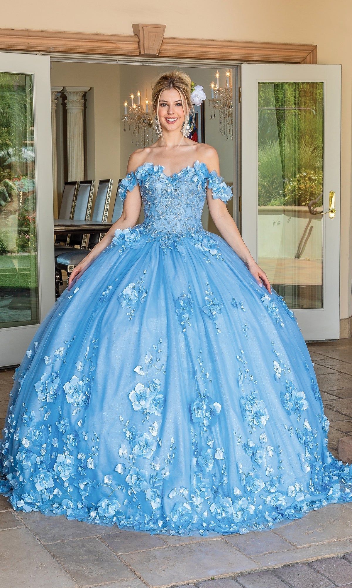 A woman smiles in front of open glass doors wearing the Quinceanera Dress 1613 By Dancing Queen, a light blue off-the-shoulder ball gown with floral embellished appliqués.