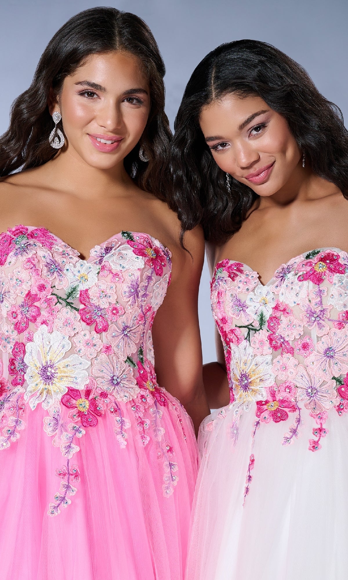 Two women stand side by side, smiling in Tiffany 16142 long prom dresses featuring strapless tulle, pink and white floral lace embroidery, and sheer corset bodices.