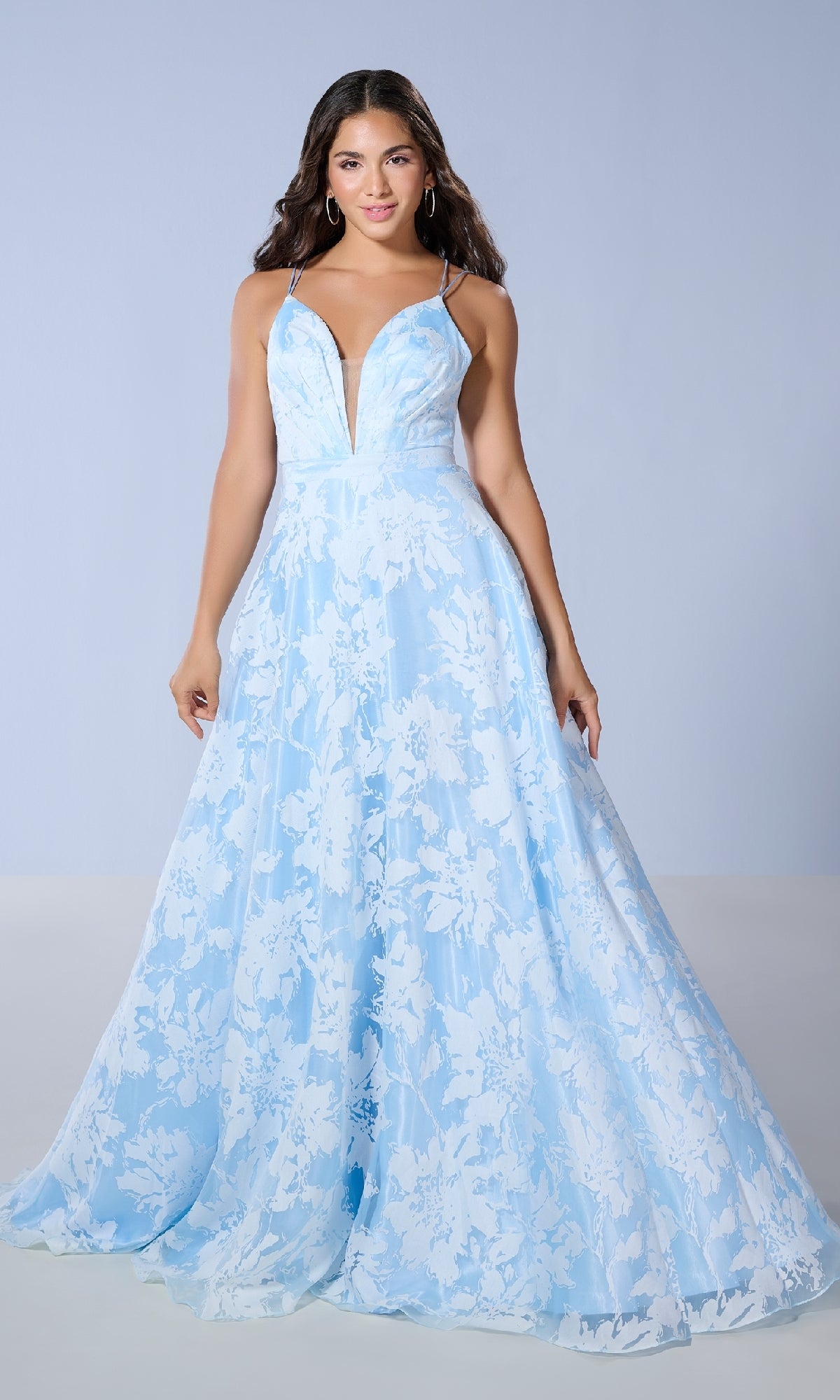 A woman wears the Tiffany 16158 long prom dress with floral print, a plunging V-neckline, and spaghetti straps in light blue and white, as she stands against a plain background.