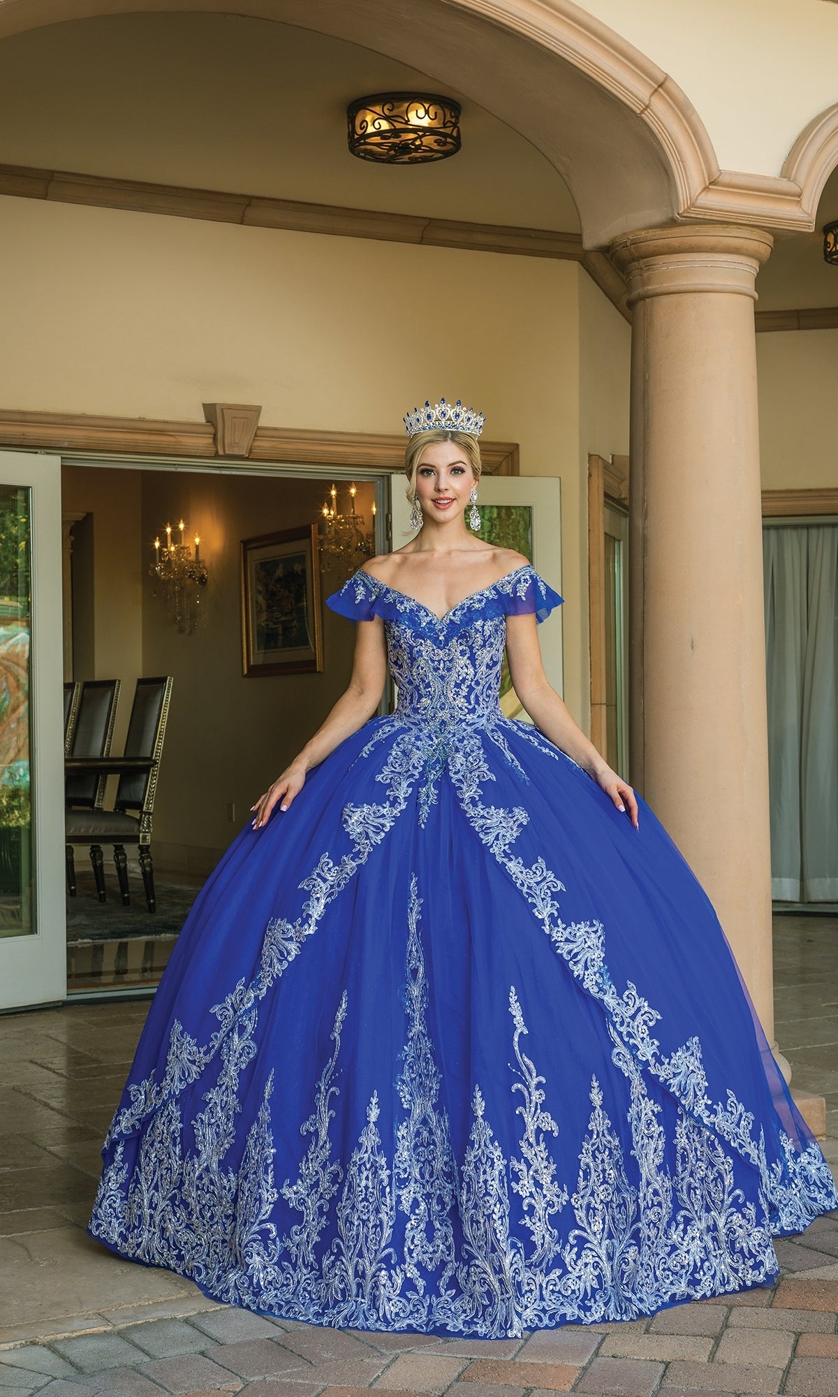 A woman in the Off-the-Shoulder Quinceanera Dress 1615, featuring blue and silver lace embroidery and a crown, stands before an open doorway with columns and gleaming chandeliers inside.