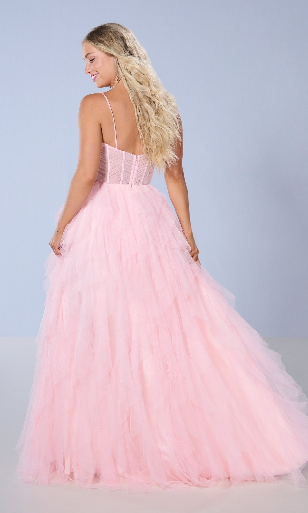 A woman with long blonde hair faces away from the camera wearing the Tiffany 16161 long prom dress, which features a light pink tulle ball gown skirt and delicate spaghetti straps, set against a plain light blue background.