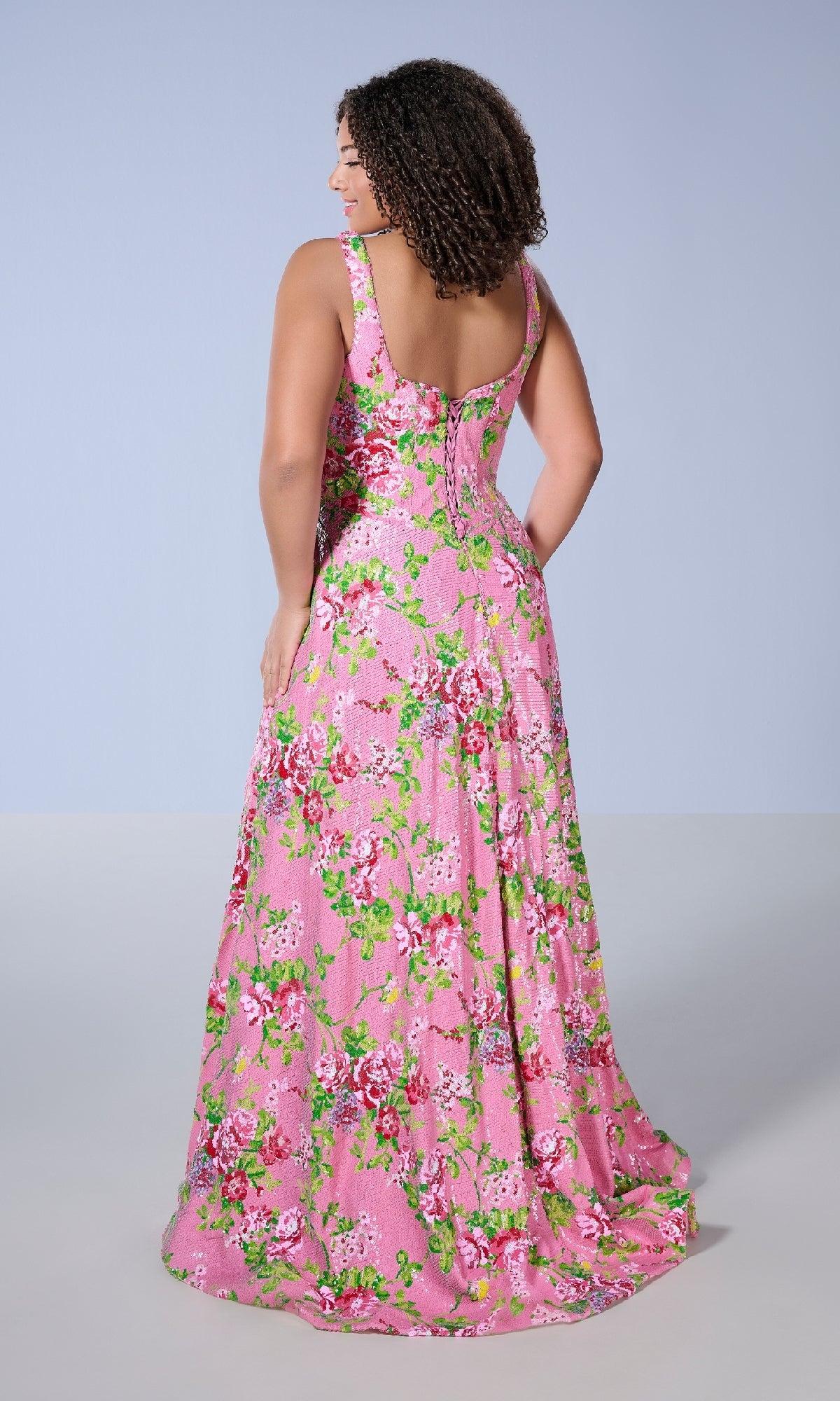 A woman faces away, wearing the Tiffany 16174 long plus-size prom dress—a sleeveless, floor-length pink gown with floral prints in green, red, and white—set against a plain background.