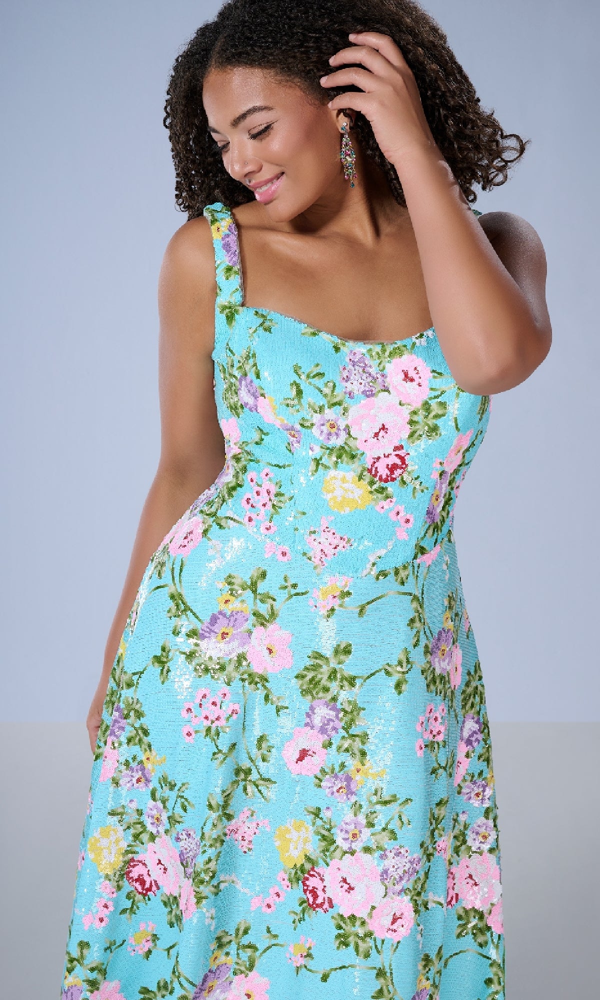 A woman poses, smiling and looking down, in the Tiffany 16174 long plus-size prom dress—a light blue floral print gown with wide straps.