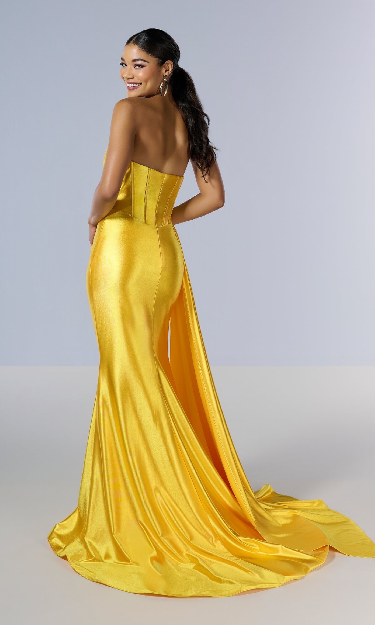 Wearing the Tiffany 16178 Long Prom Dress—a strapless, form-fitting yellow satin gown with a corset bodice and long train—a woman stands facing away, smiling over her shoulder.