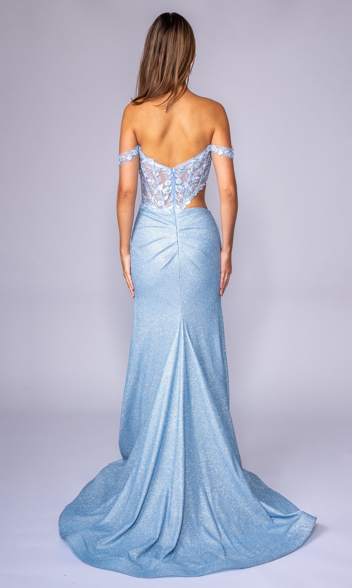 A woman wears the Nina Canacci 1618, a long formal light blue off-the-shoulder prom dress with lace, sheer back detailing, and delicate floral embroidery, posing elegantly against a plain background.