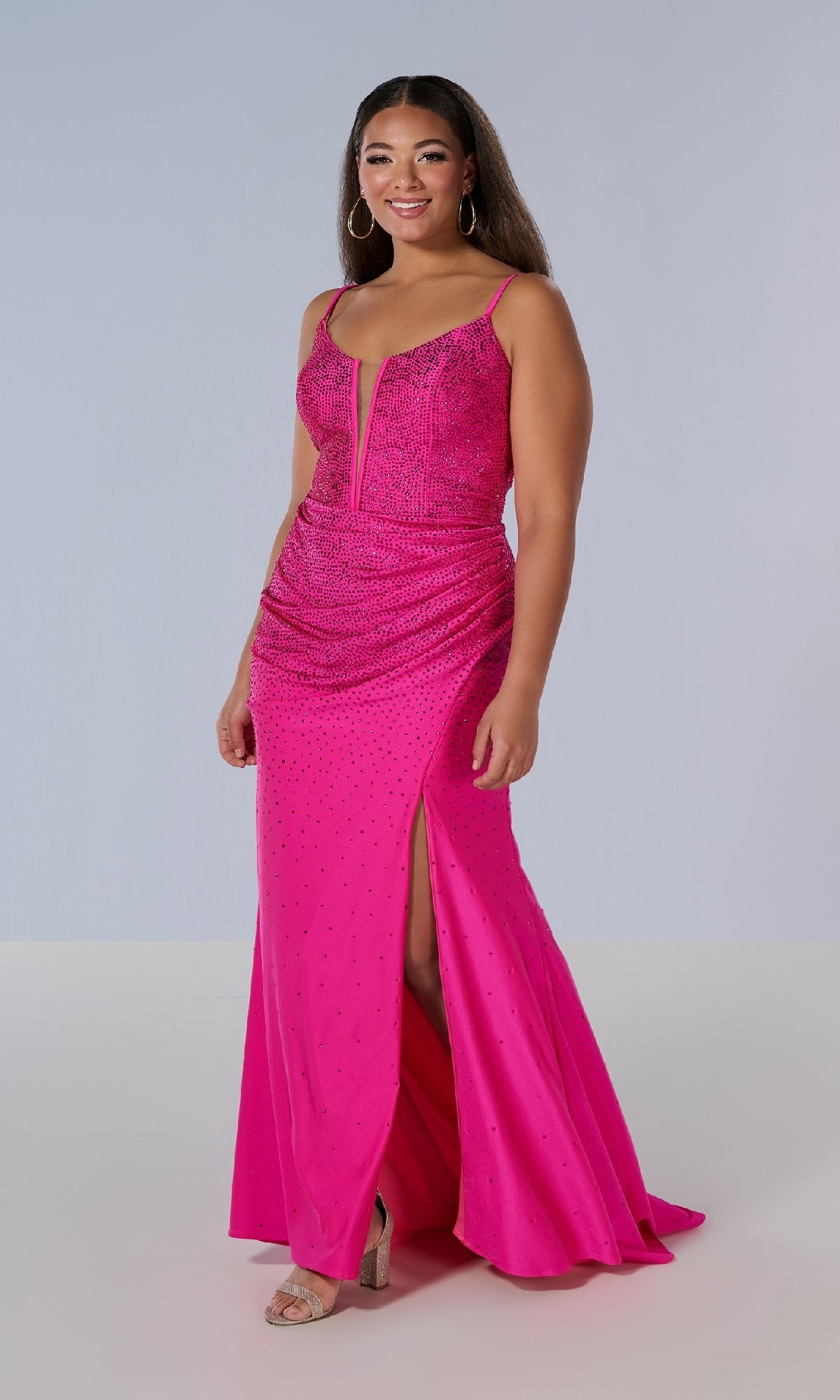 A woman wears the Tiffany 16194 long plus-size prom dress featuring a bright pink hue, corset bodice, deep V neckline, ruched waist, beaded details, and a thigh-high slit, standing against a light background.