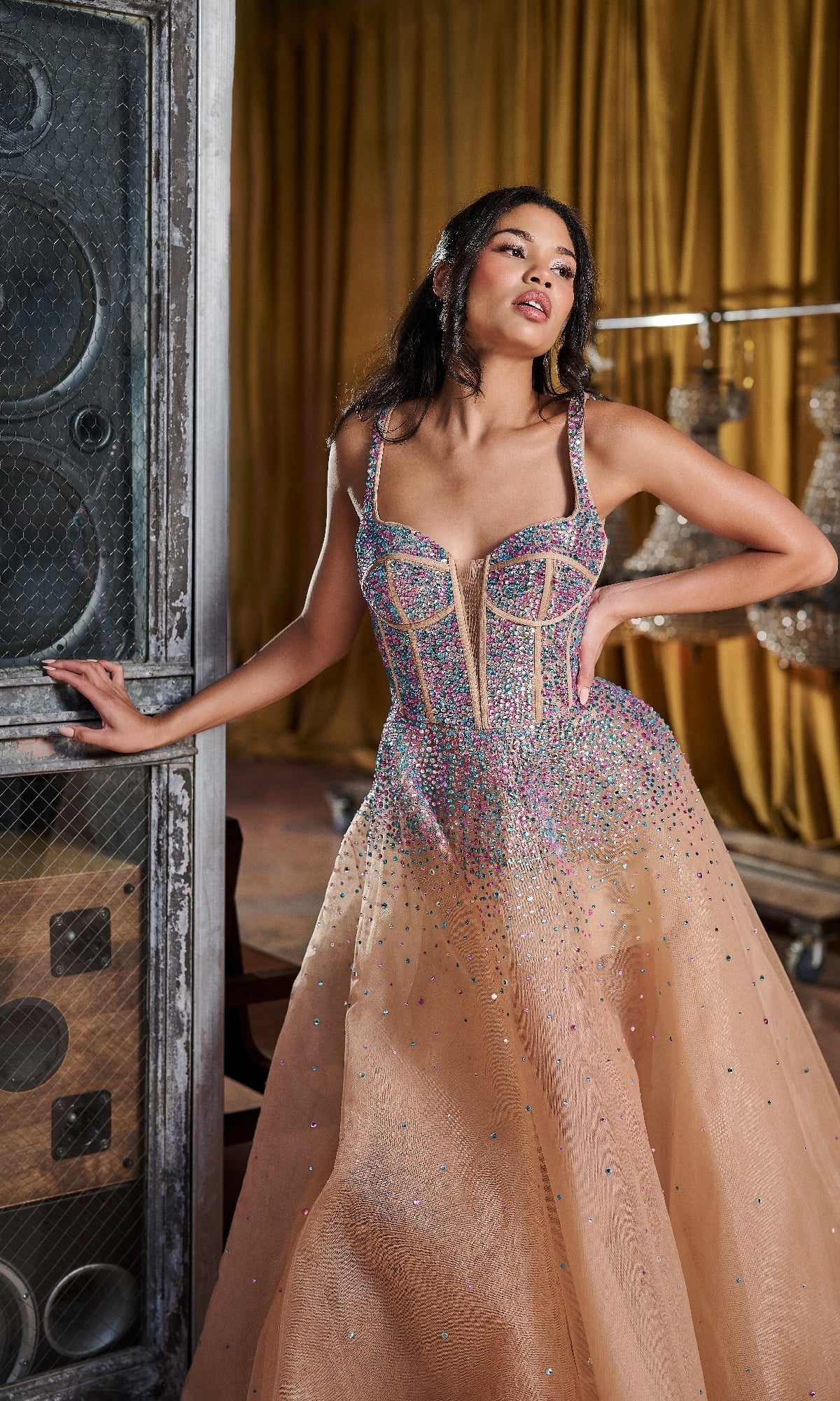 A woman in the Long Prom Dress: Tiffany 16206, featuring sparkly beaded tulle and crystal embellishments, poses indoors beside a large speaker and draped gold curtains.