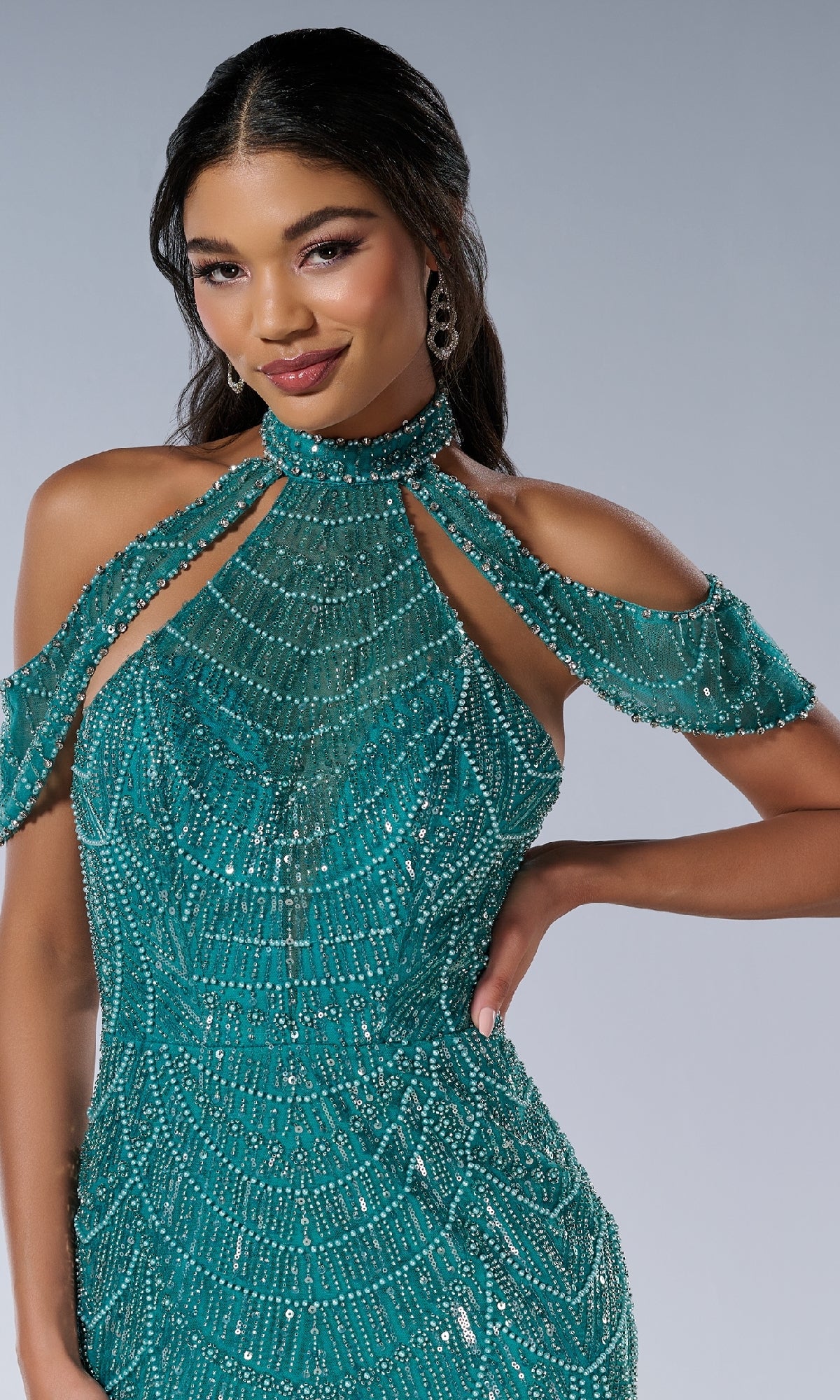 A woman poses with one hand on her hip in the Tiffany 16207— a fitted, floor-length teal prom dress featuring beaded details, a halter neckline, and cold-shoulder sleeves against a plain background.
