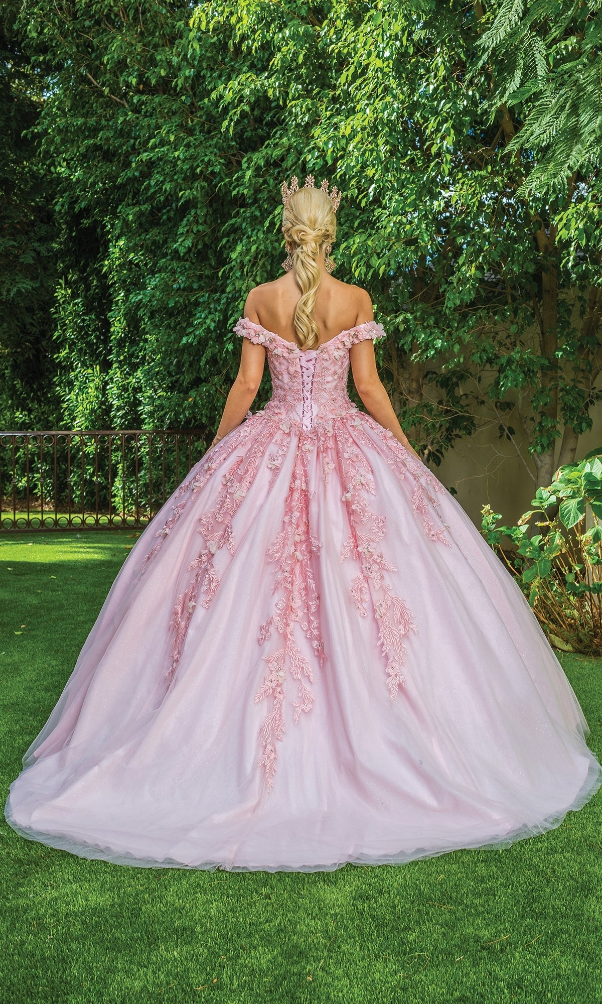 A woman wearing the Quinceanera Dress 1620 By Dancing Queen, a light pink off-the-shoulder gown with lace details and a crown, stands on grass facing away, with trees in the background.