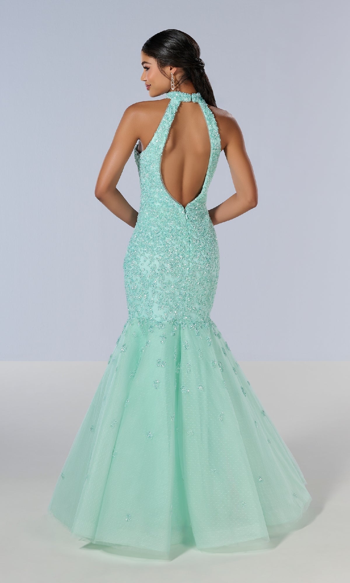 A woman wearing the Tiffany 16210 long prom dress—a light blue, beaded tulle mermaid gown with a halter neckline and open back—stands against a plain background.
