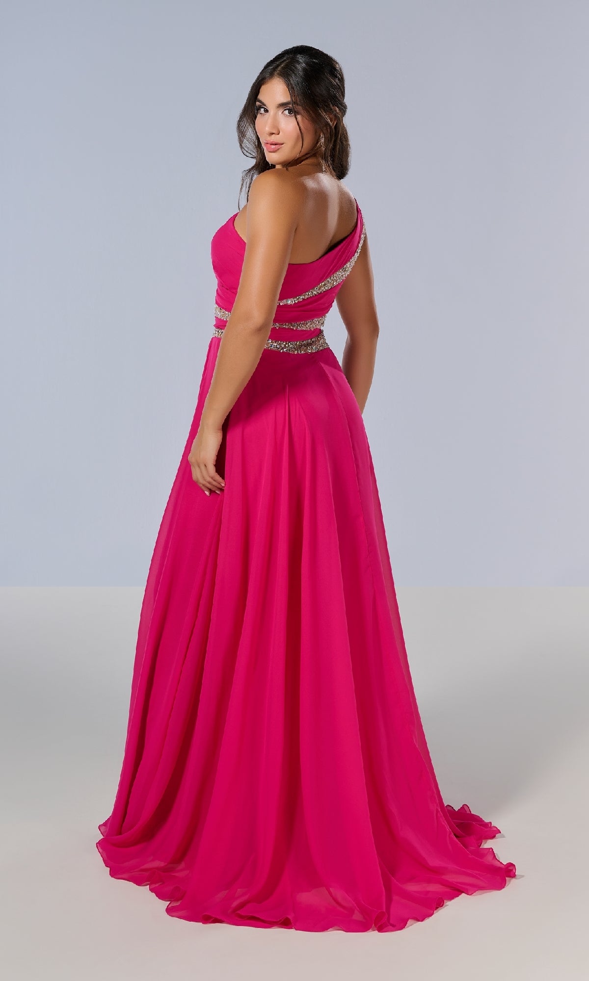 A woman in the Tiffany 16217 long prom dress, a bright pink, floor-length one-shoulder gown with silver embellishments, stands facing away and looks over her shoulder.