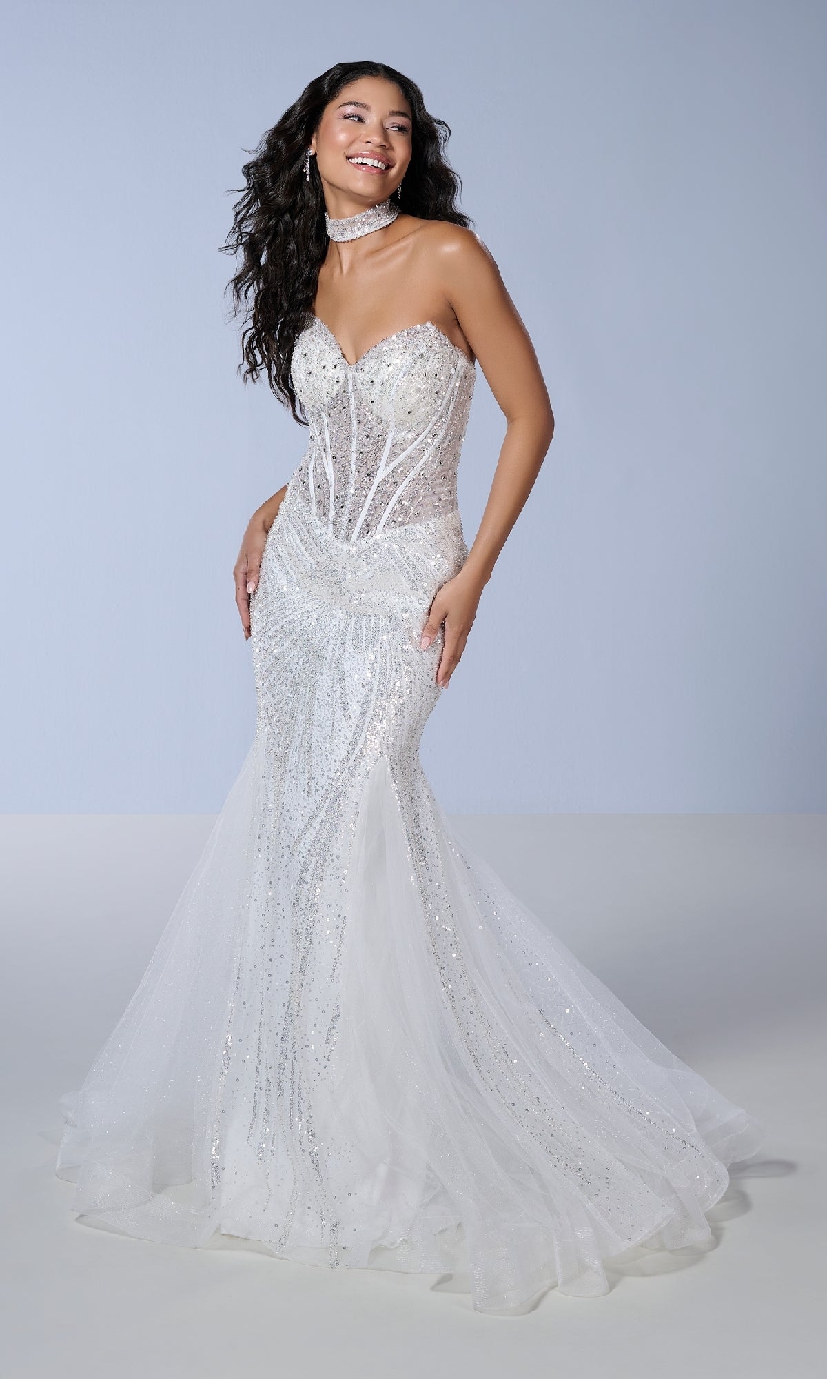 A woman poses against a plain light background wearing the Tiffany 16232 long prom dress—a strapless mermaid gown with a sweetheart neckline, sparkling sequin-embellished tulle, and a sheer overlay.