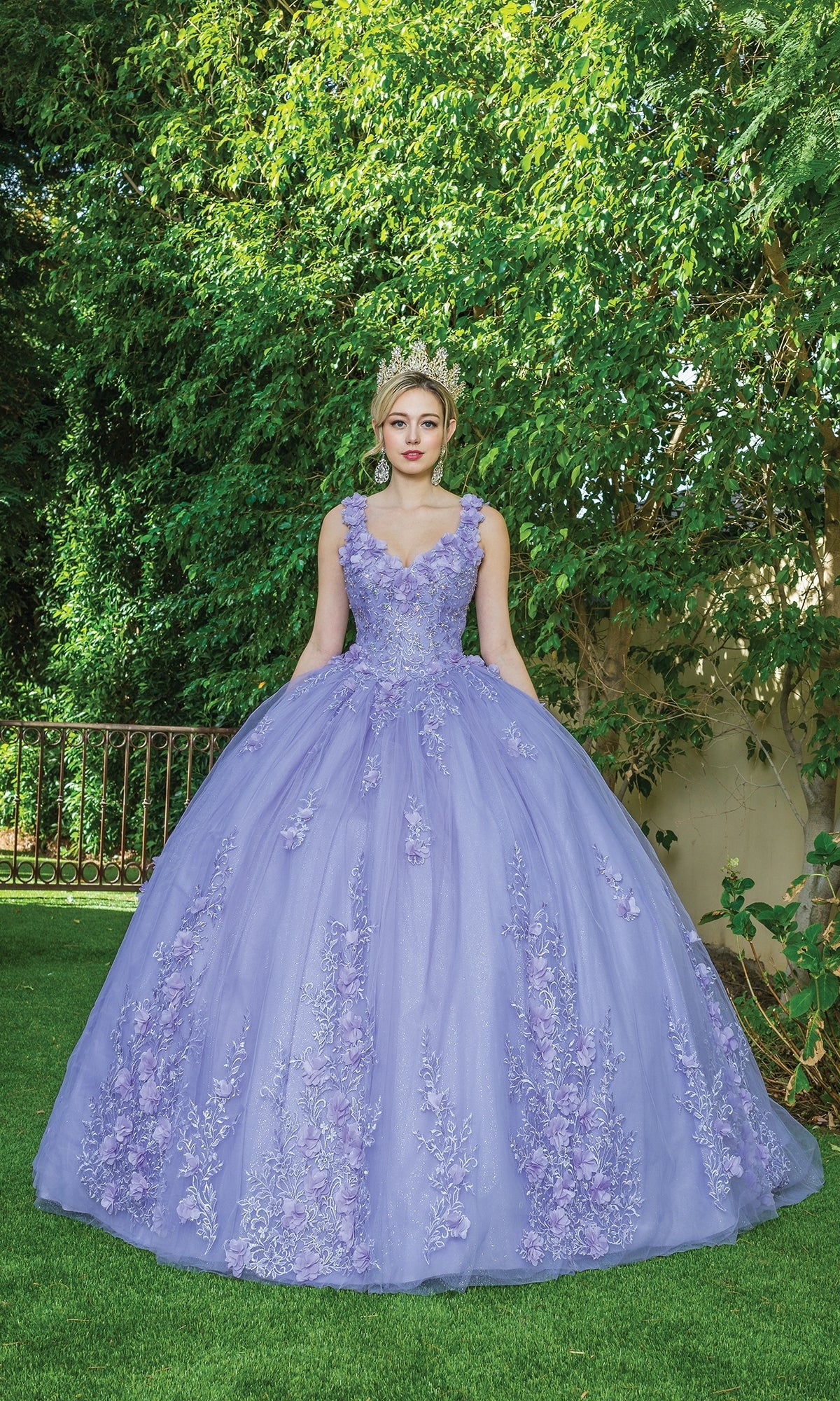 A person wearing the Quinceanera Dress 1623 By Dancing Queen and a tiara stands on grass in front of a green leafy background.
