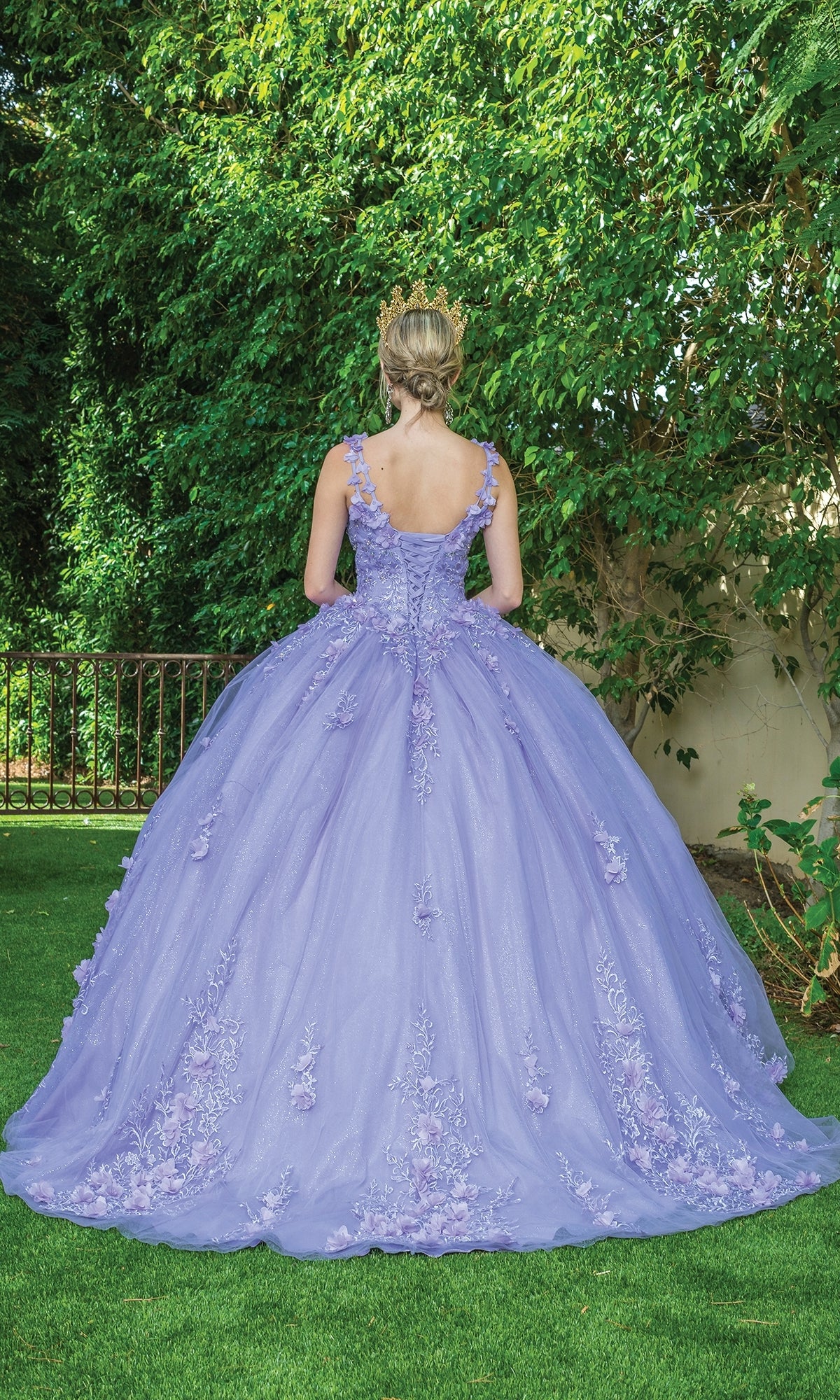 A woman in the Quinceanera Dress 1623 by Dancing Queen and a tiara stands on grass with her back to the camera, facing a lush, leafy green background.