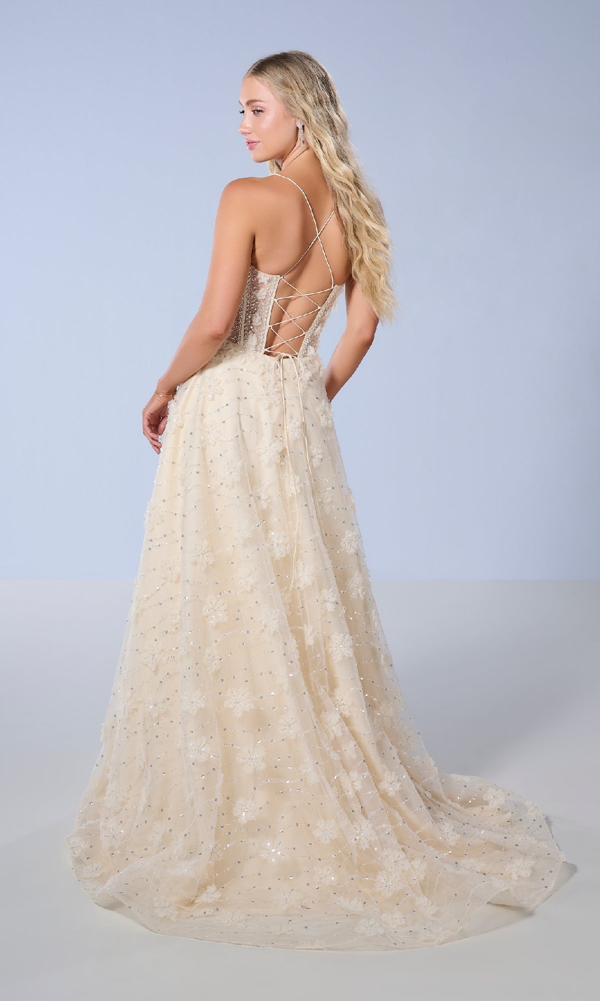 A woman poses against a plain, light background wearing the Long Prom Dress: Tiffany 16247—a floor-length champagne gown featuring a sheer bodice, sequin accents, and a delicate floral pattern.