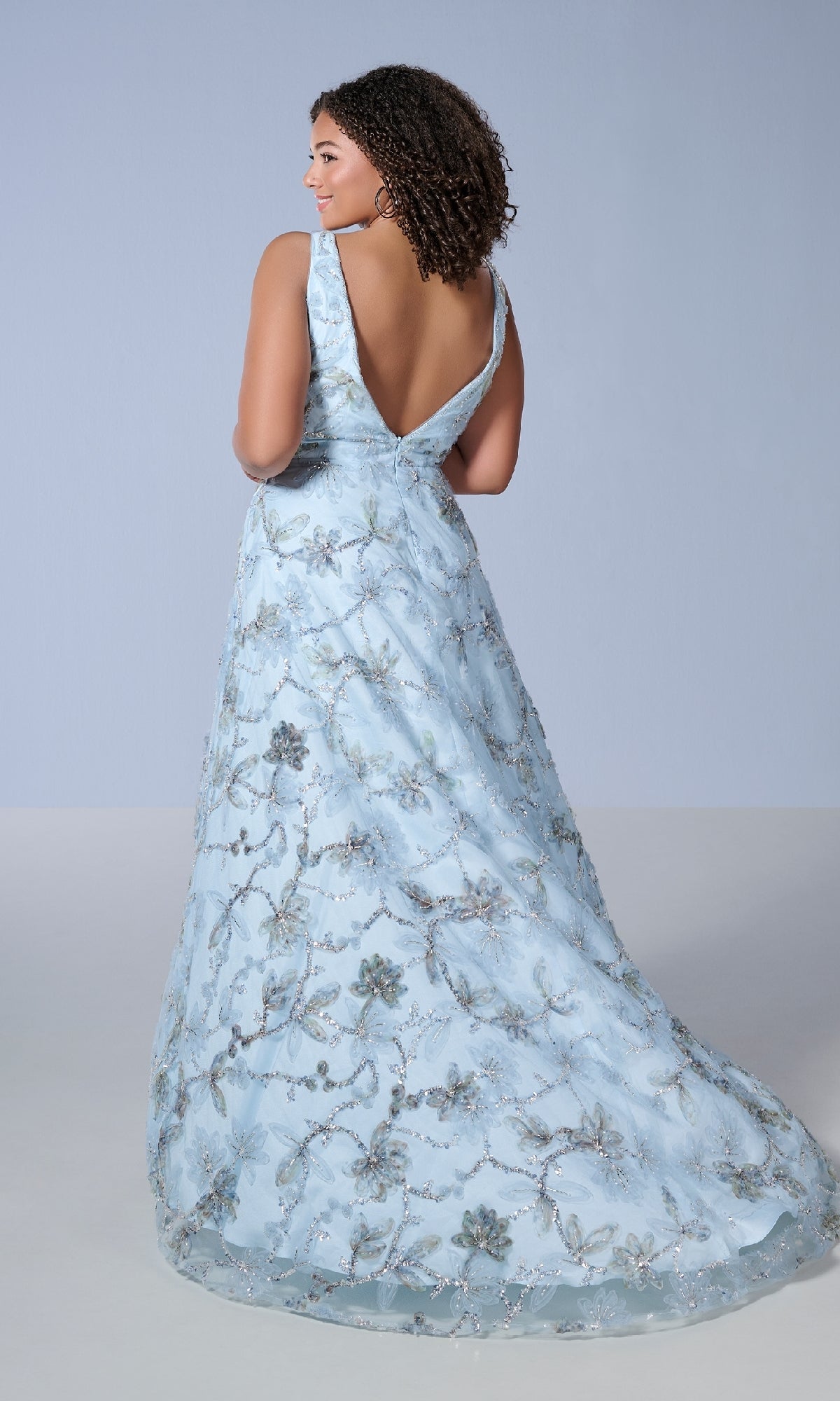 A woman showcases the Long Plus-Size Prom Dress Tiffany 16249, featuring light blue fabric with silver floral embroidery, a deep V-back, and elegant embellished details.