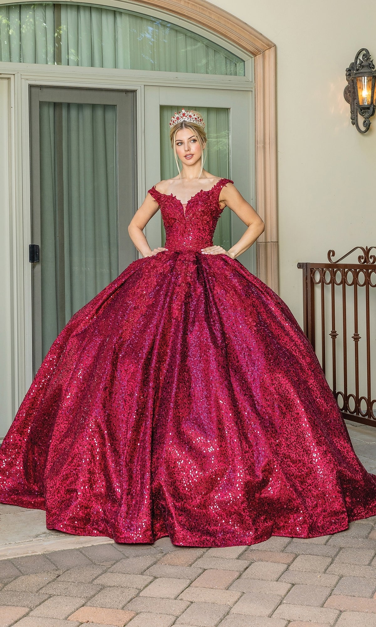 A woman in the Quinceanera Dress 1624 By Dancing Queen, a sparkling red off-the-shoulder gown with a sweetheart neckline and tiara, stands outdoors on a stone patio before glass doors and an iron railing.