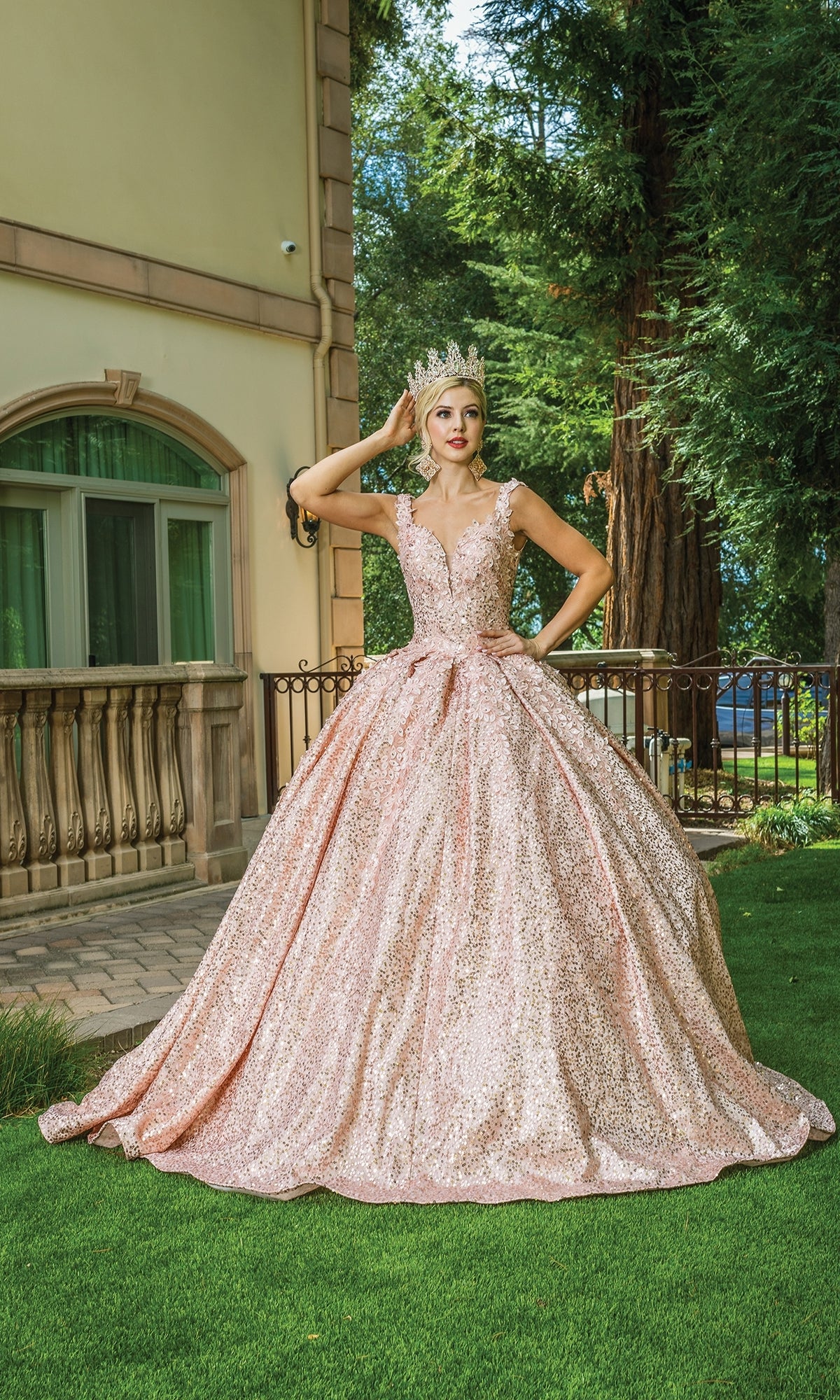 A woman wearing the Quinceanera Dress 1624 By Dancing Queen—a sparkling pink off-shoulder gown with a sweetheart neckline—poses on a lawn before a large building and tall trees, her crown gleaming in the sunlight.