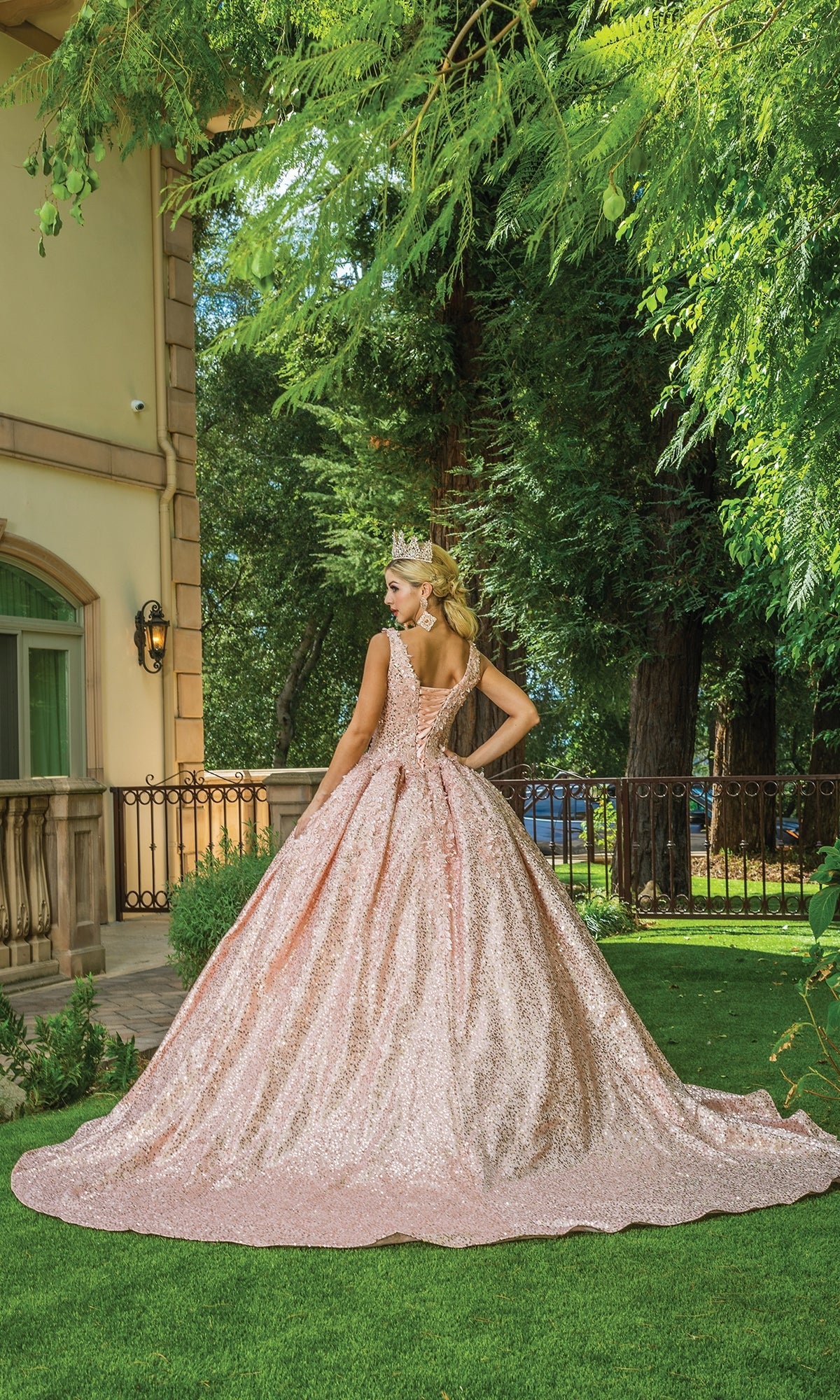A woman in the Quinceanera Dress 1624 By Dancing Queen, a sparkling light pink off-the-shoulder gown, stands outdoors on green grass facing away near a building and tall trees, wearing a tiara.