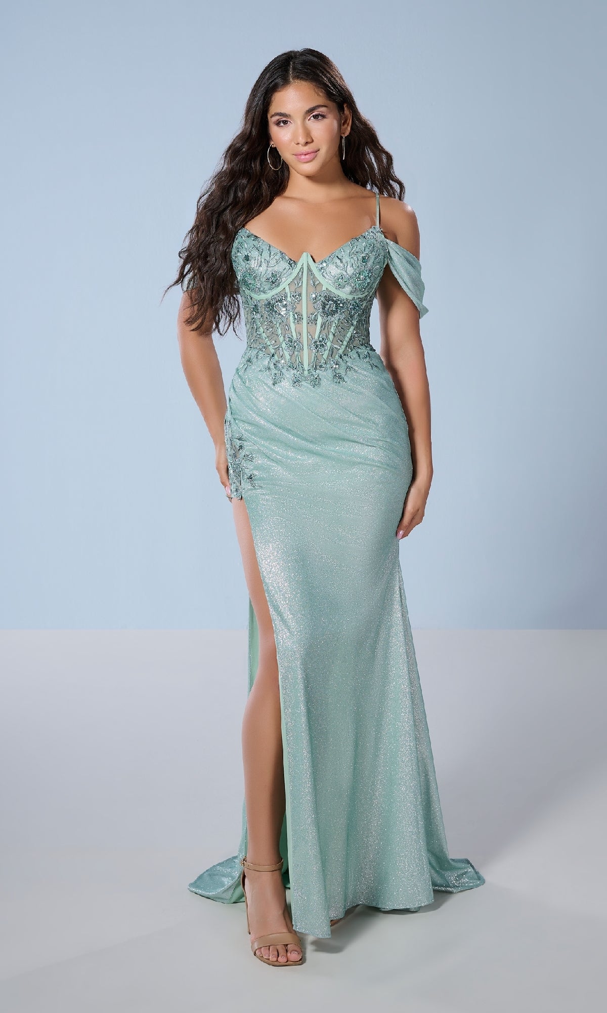 A woman in the Tiffany 16250 long prom dress—a shimmery mint green gown with rhinestones, spaghetti straps, lace embroidery, and side slit—stands in open-toe heels against a light blue background.