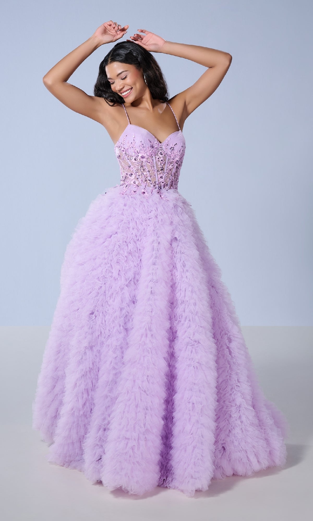 A woman poses with arms raised in the Long Prom Dress: Tiffany 16252, featuring a light purple ruffled tulle skirt, beaded bodice, and lace appliques against a plain background.