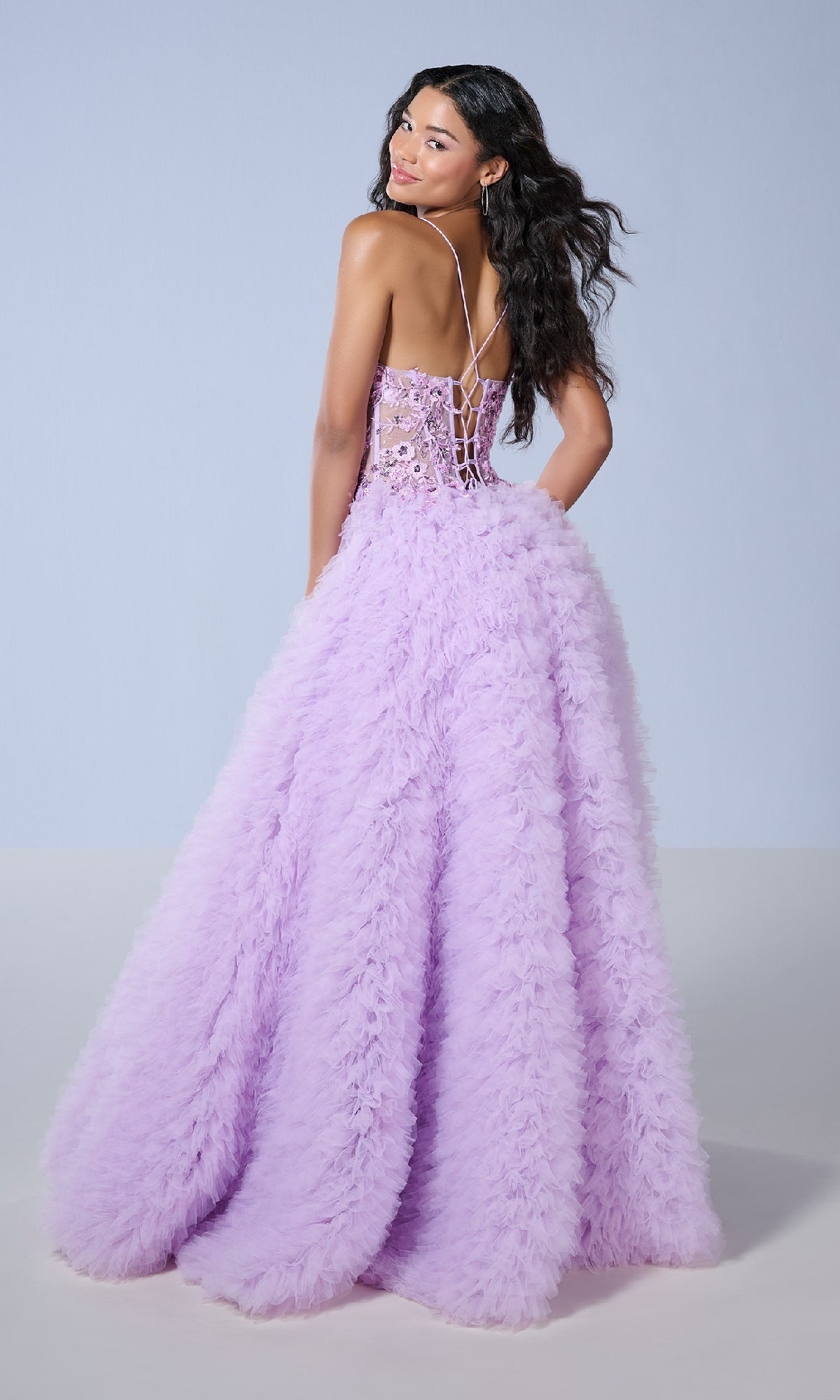 A woman in the Tiffany 16252 long prom dress—backless, lavender tulle with ruffles, lace-up corset bodice, and lace appliqués—stands against a plain background, glancing over her shoulder.