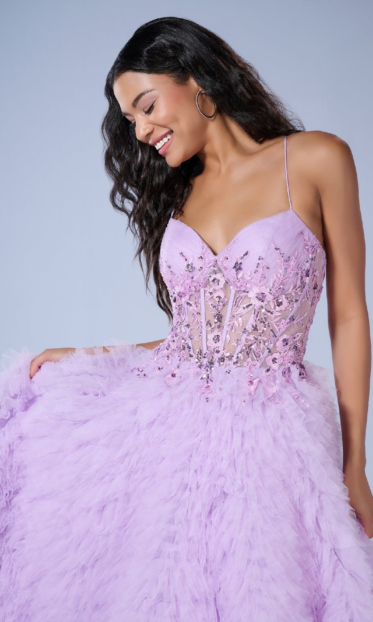 A woman smiles against a plain background wearing the Long Prom Dress: Tiffany 16252, featuring a lavender corset bodice with beaded details and a feathered skirt as she holds out the dress.