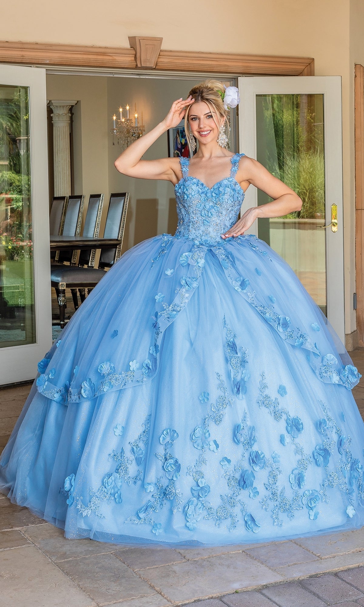 A woman stands outside double doors wearing the Quinceanera Dress 1627 By Dancing Queen, a light blue ball gown with floral details, a full tulle skirt, and a graceful sweetheart neckline.