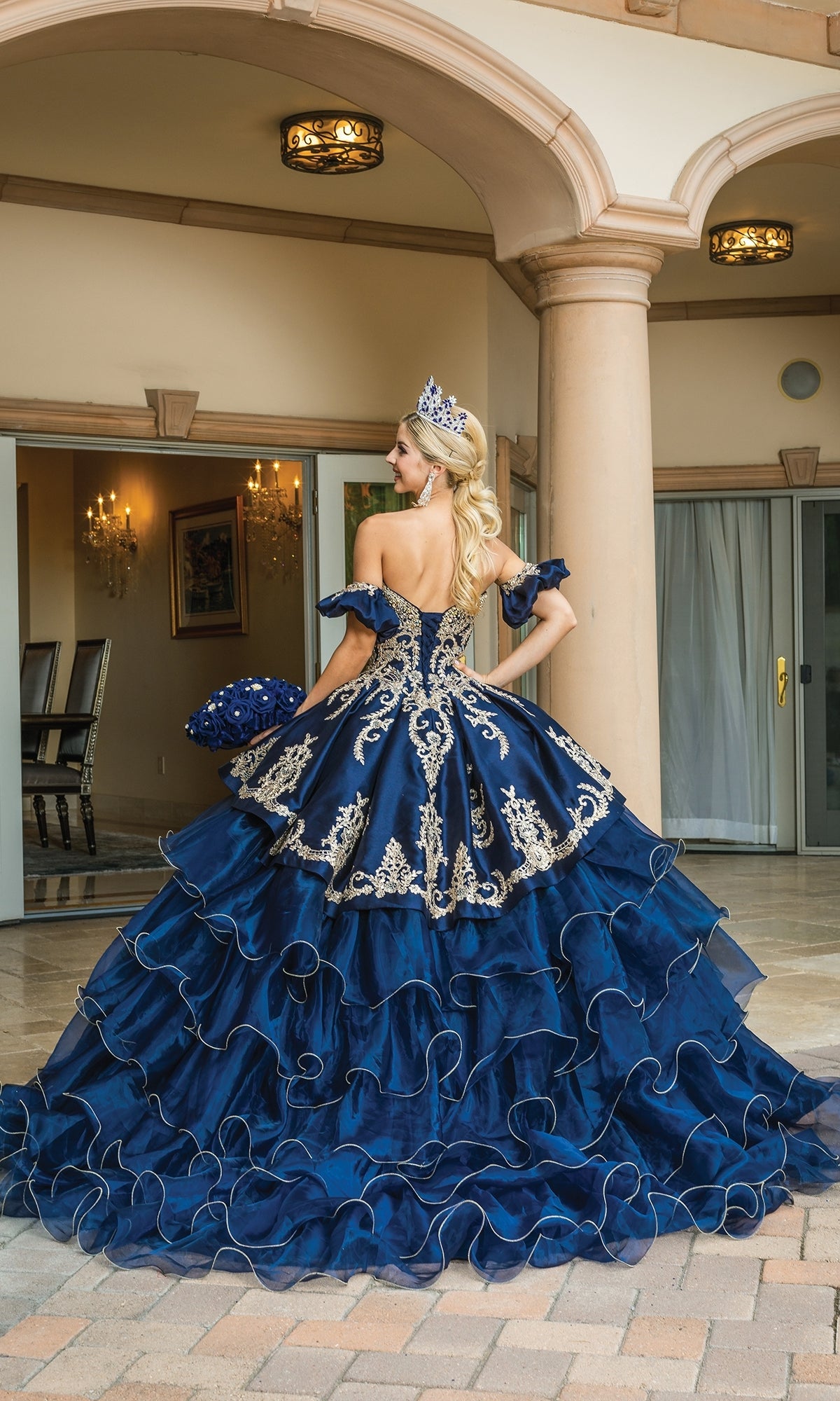 A woman wearing the Navy Blue Quinceanera Dress 1637, featuring gold embroidery, a ruffled layered skirt, and a tiara, stands outside a building with her back to the camera.