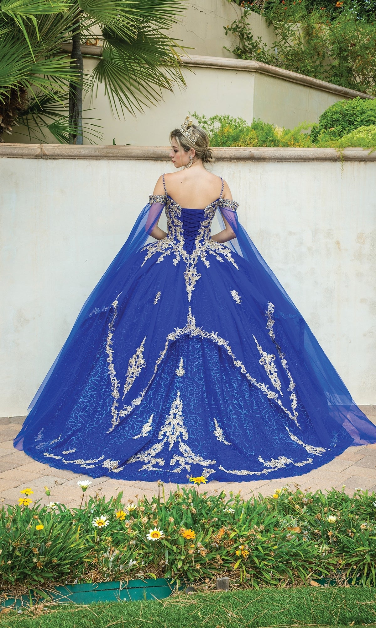 A woman stands outdoors in greenery and yellow flowers, wearing the Quinceanera Dress 1638 By Dancing Queen—an elaborate off-the-shoulder blue ball gown with silver embroidery and a tiara, facing a wall.