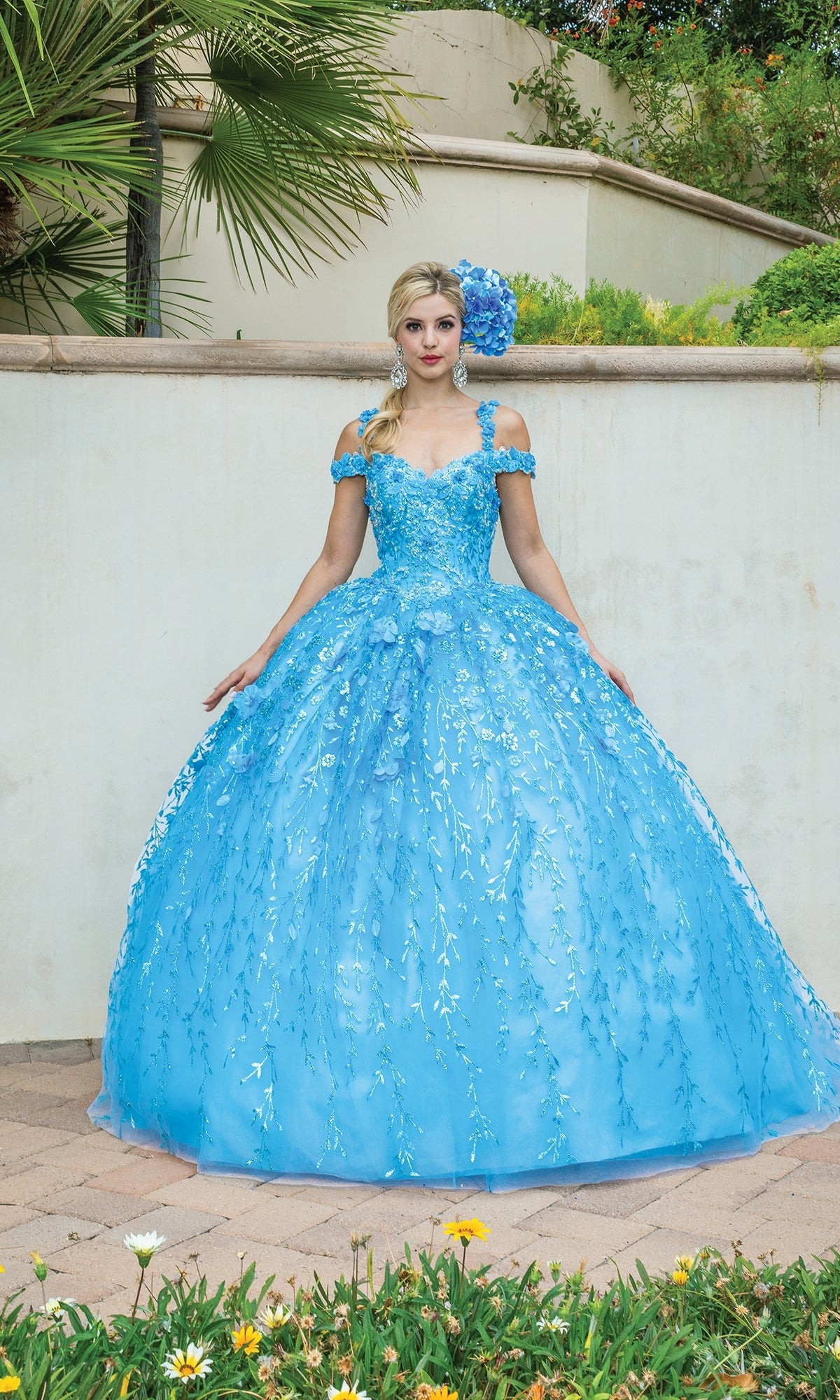 A woman stands outdoors in front of a white wall, wearing the Quinceanera Dress 1640 by Dancing Queen—an off-the-shoulder A-line ball gown with bright blue floral details and a matching floral headpiece.