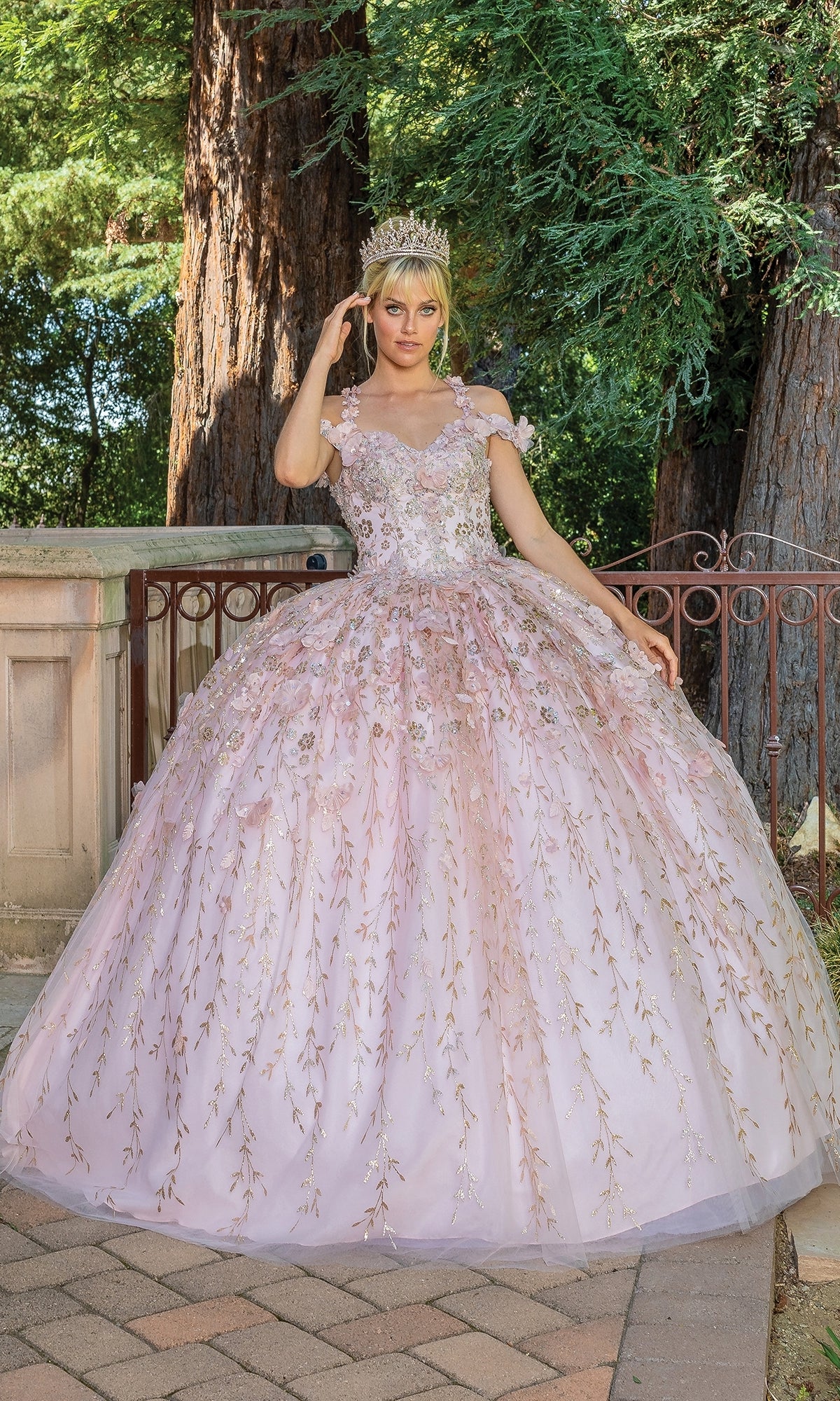 A person stands outdoors on a paved area, wearing the Quinceanera Dress 1640 By Dancing Queen—an off-the-shoulder pink a-line ball gown with floral details and a crown, with trees and a railing in the background.