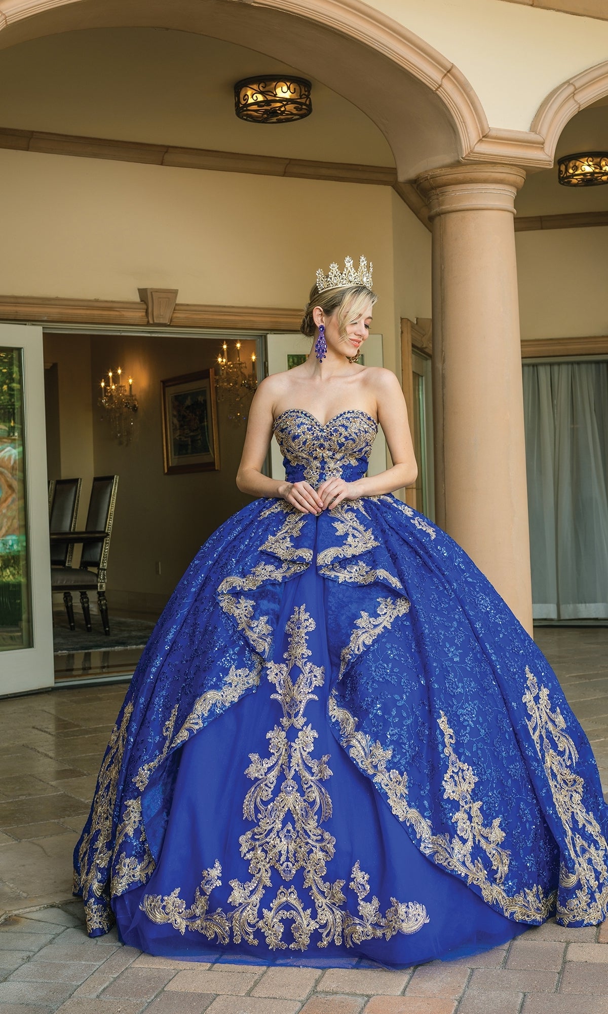 A woman in the Quinceanera Dress 1650 By Dancing Queen, a blue and gold ornate ball gown with gold lace embroidery, crown, and earrings, stands outside a building with columns and an arched entryway.