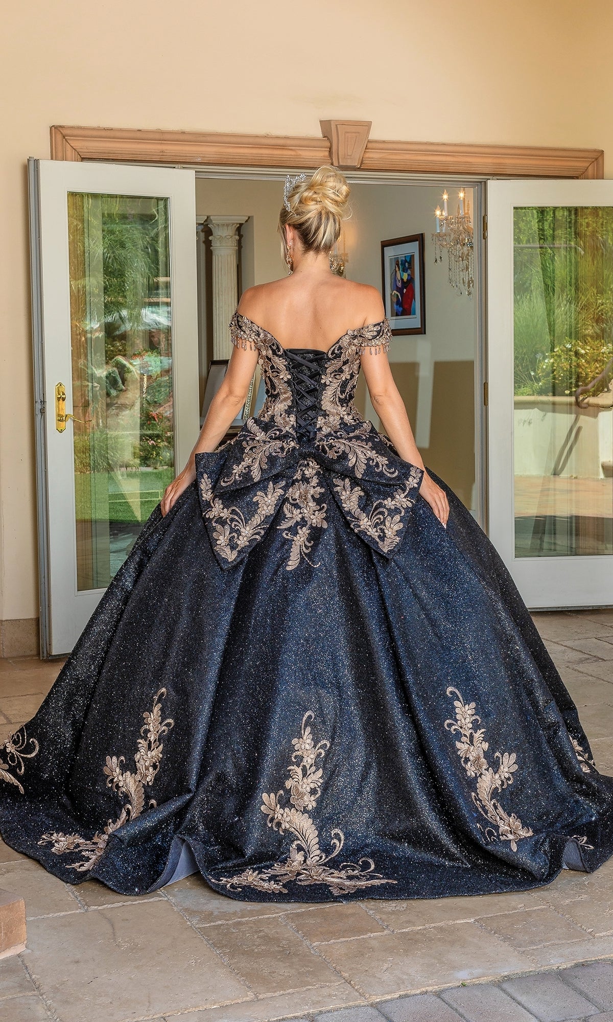 A woman in an off-the-shoulder Quinceanera Dress 1658 By Dancing Queen stands facing away before open double doors, her dark blue skirt featuring intricate gold embroidery.