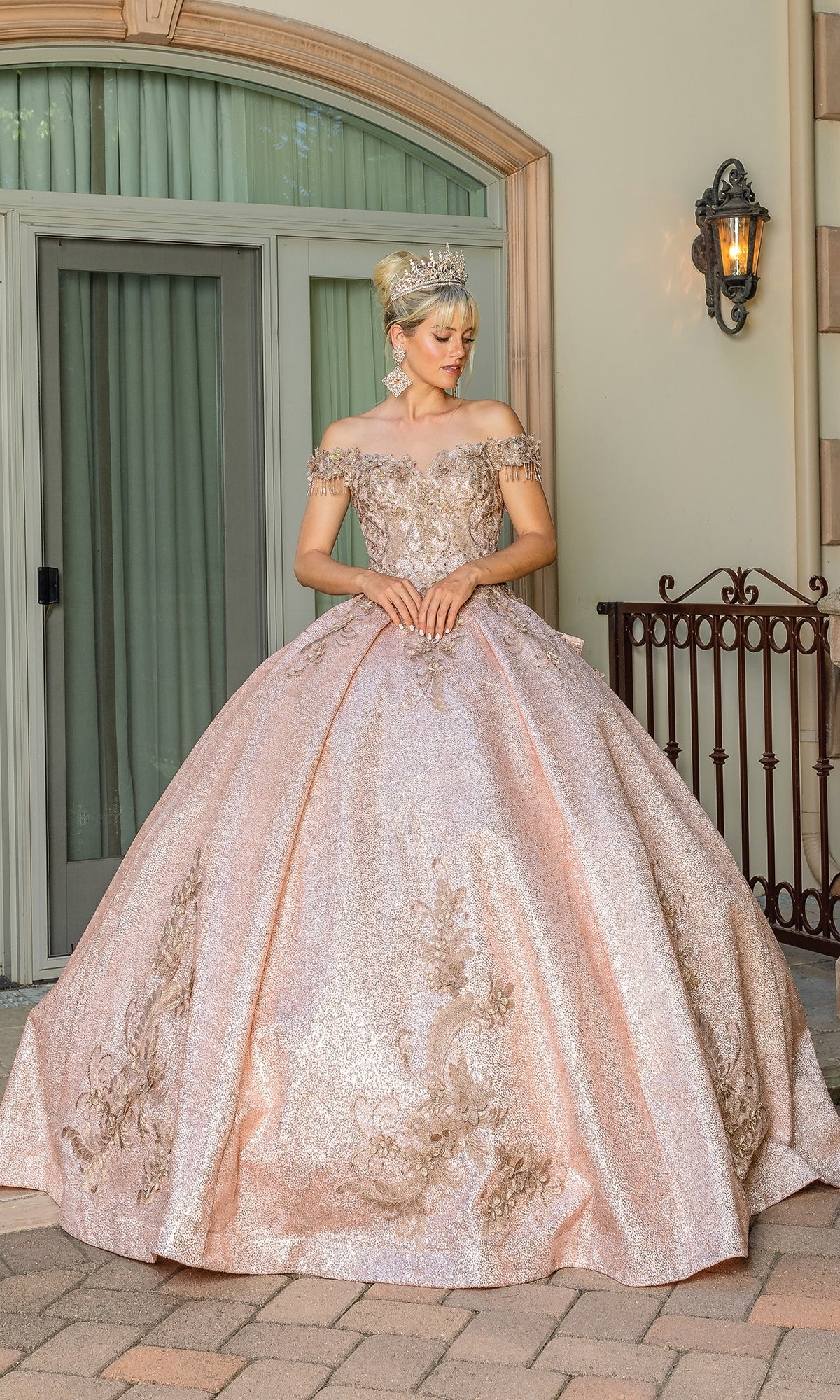 A woman stands outdoors wearing the Quinceanera Dress 1658 by Dancing Queen, an ornate light pink off-the-shoulder ball gown with gold embellishments and a tiara, perfect for a royal celebration.
