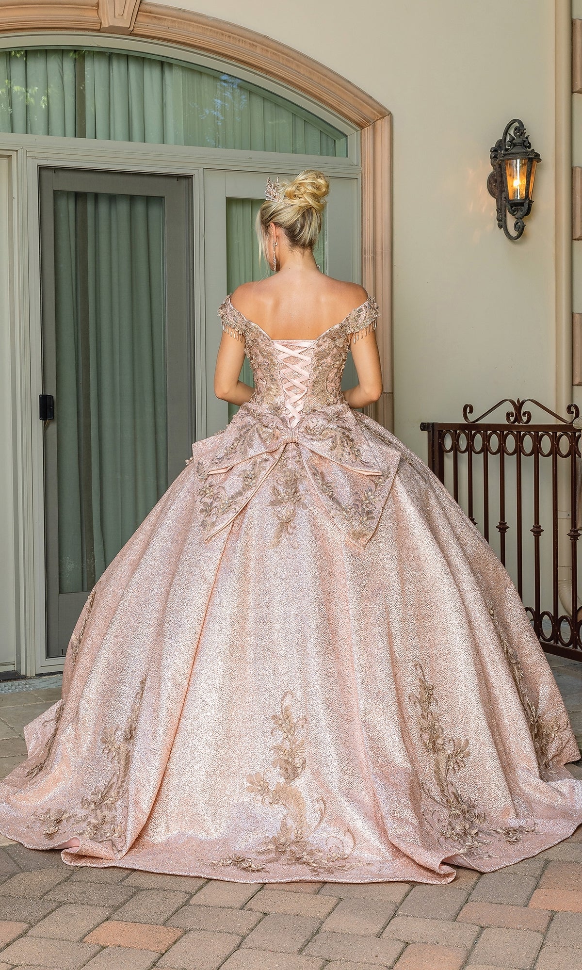 A woman stands outdoors in the Quinceanera Dress 1658 By Dancing Queen, featuring an off-the-shoulder design, shimmering pink A-line skirt, gold embellishments, and a large bow as she faces away from the camera.