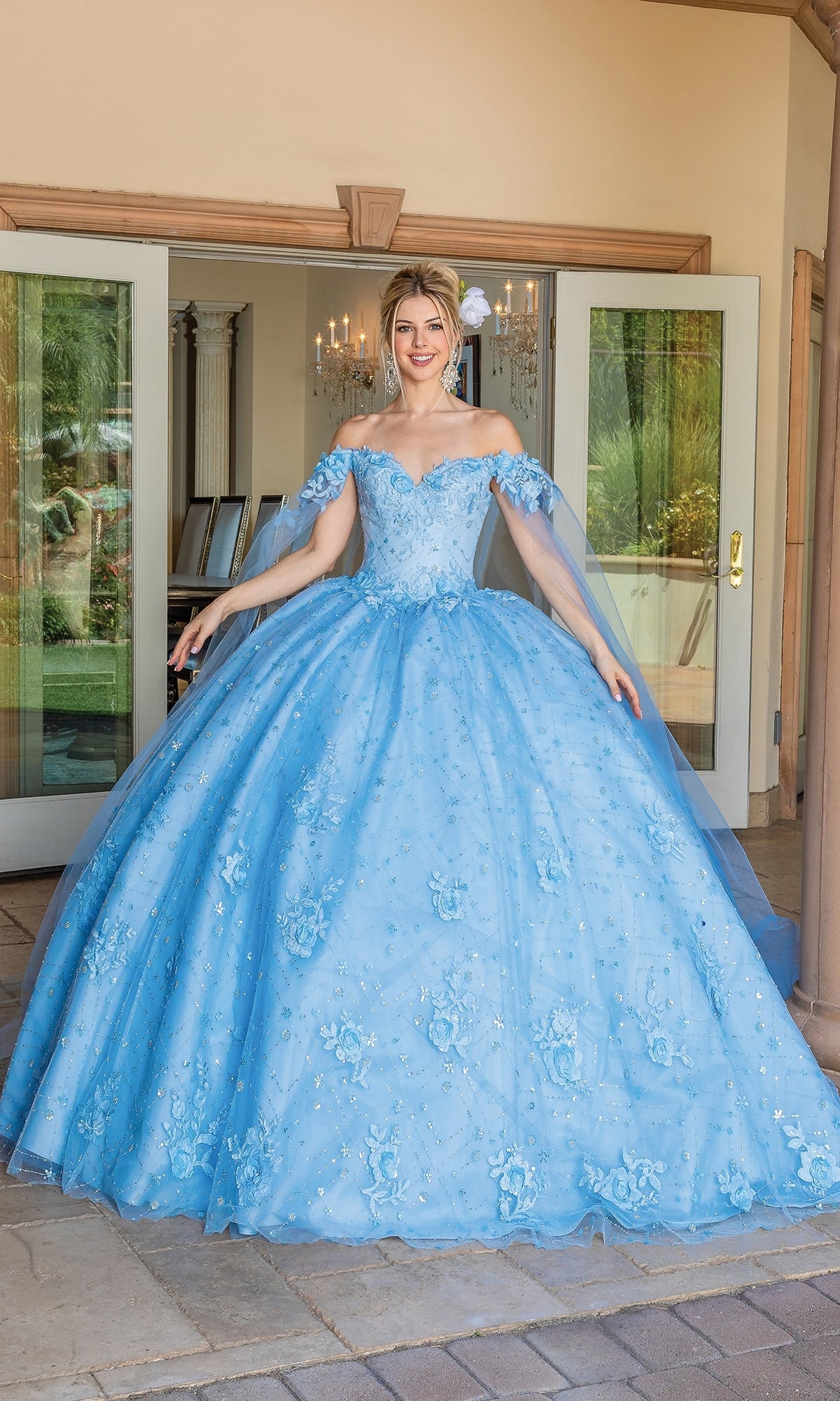 A woman stands outside double doors in the Quinceanera Dress 1660 By Dancing Queen—a light blue, off-the-shoulder gown with floral embellishments, full tulle skirt, and delicate removable cape sleeves.